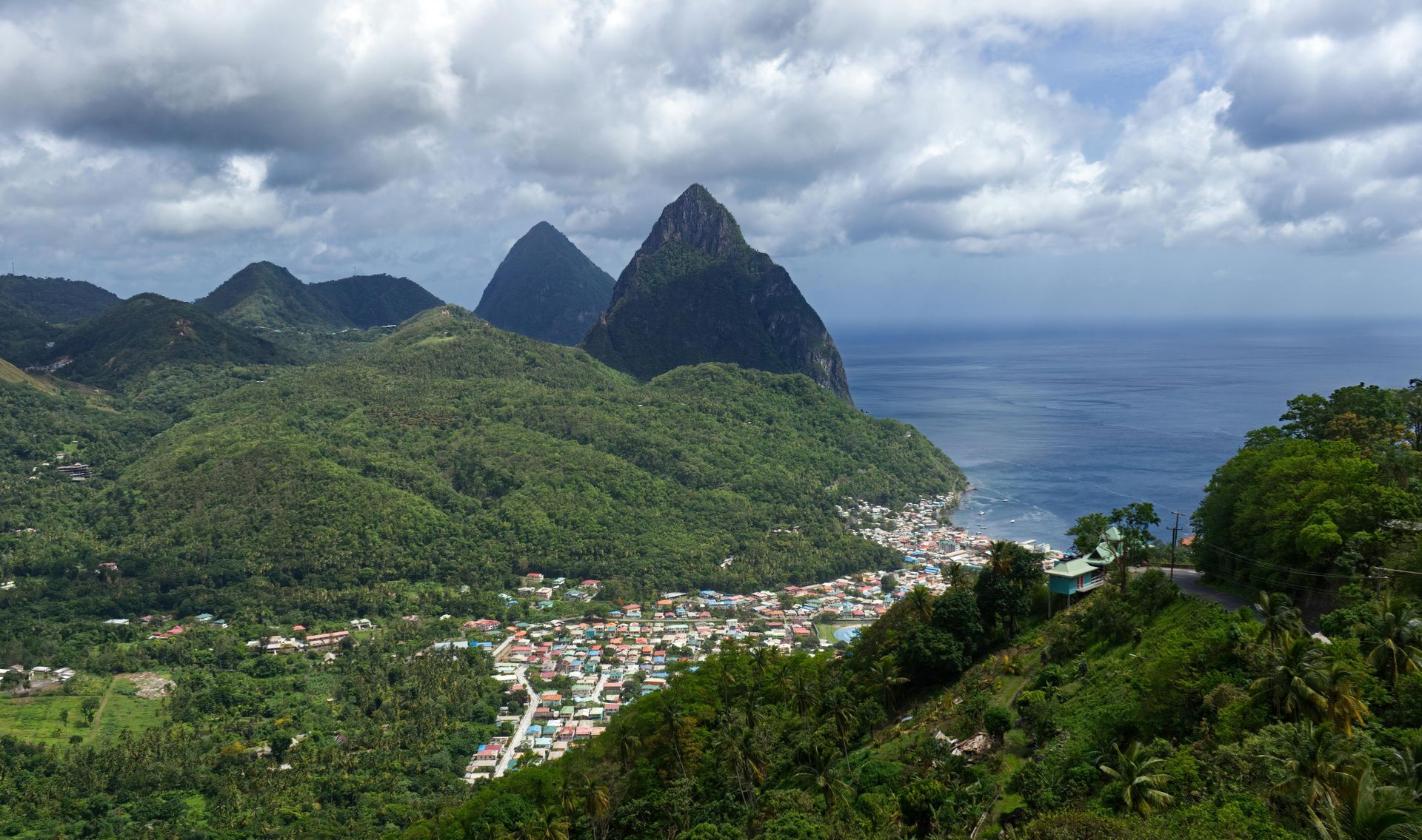 Green mountains overlook a town on the coast, with the Pitons and ocean in the distance under a cloudy sky.