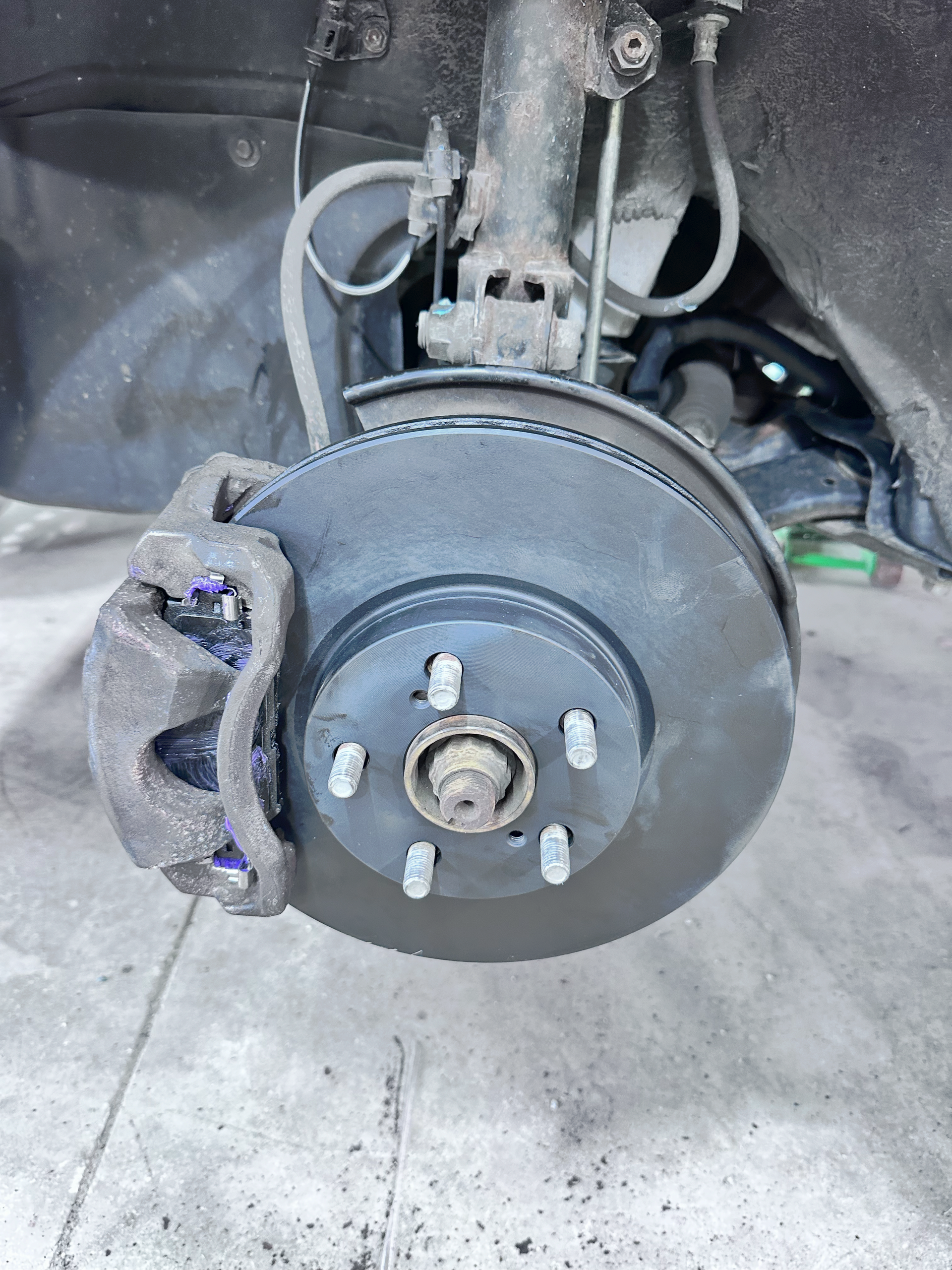 Car's front brake assembly: rotor, caliper, and suspension components, viewed up close. | Midwest Tire & Service LLC