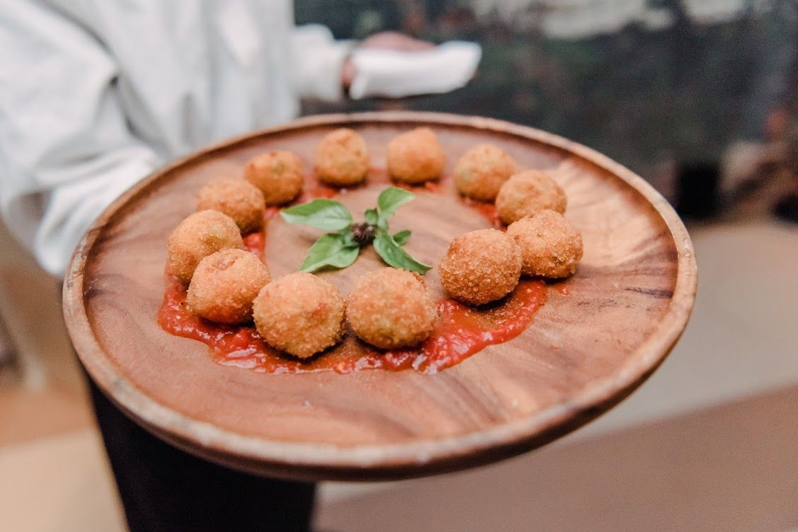 A person holding a wooden plate of fried balls with red sauce and basil.