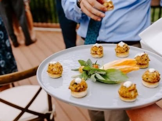 Person holding a plate of appetizers for catering event in Annapolis, MD on a wooden deck. The plate has crab cakes topped with sauce, herbs, and yellow flowers.