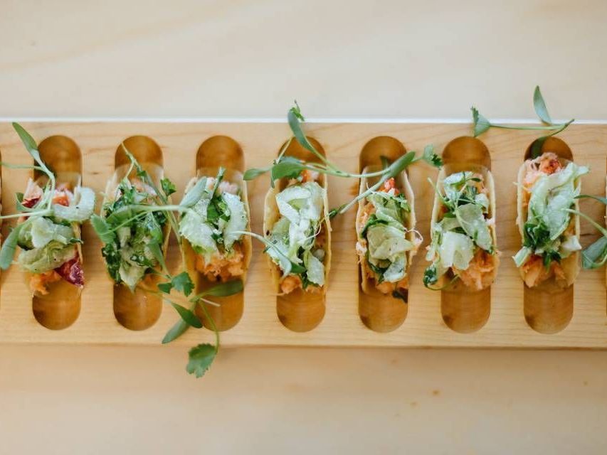 Tacos in small wooden holders, topped with greens, arranged on a wooden serving board.