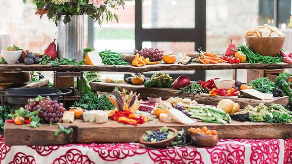 Buffet table with various fruits, vegetables, cheeses, and dips, set on wooden planks over a patterned tablecloth.