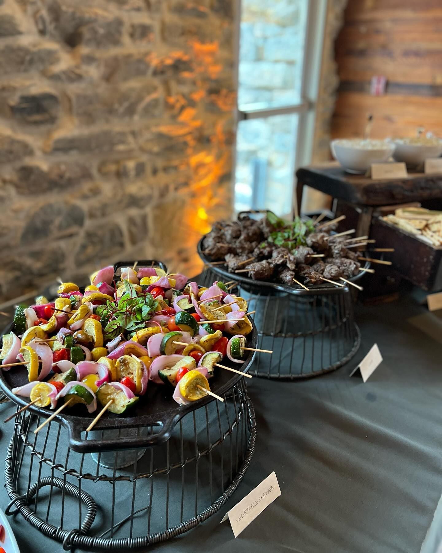 Lunch Buffet in M: grilled vegetable and meat skewers on a table, with a stone wall backdrop.