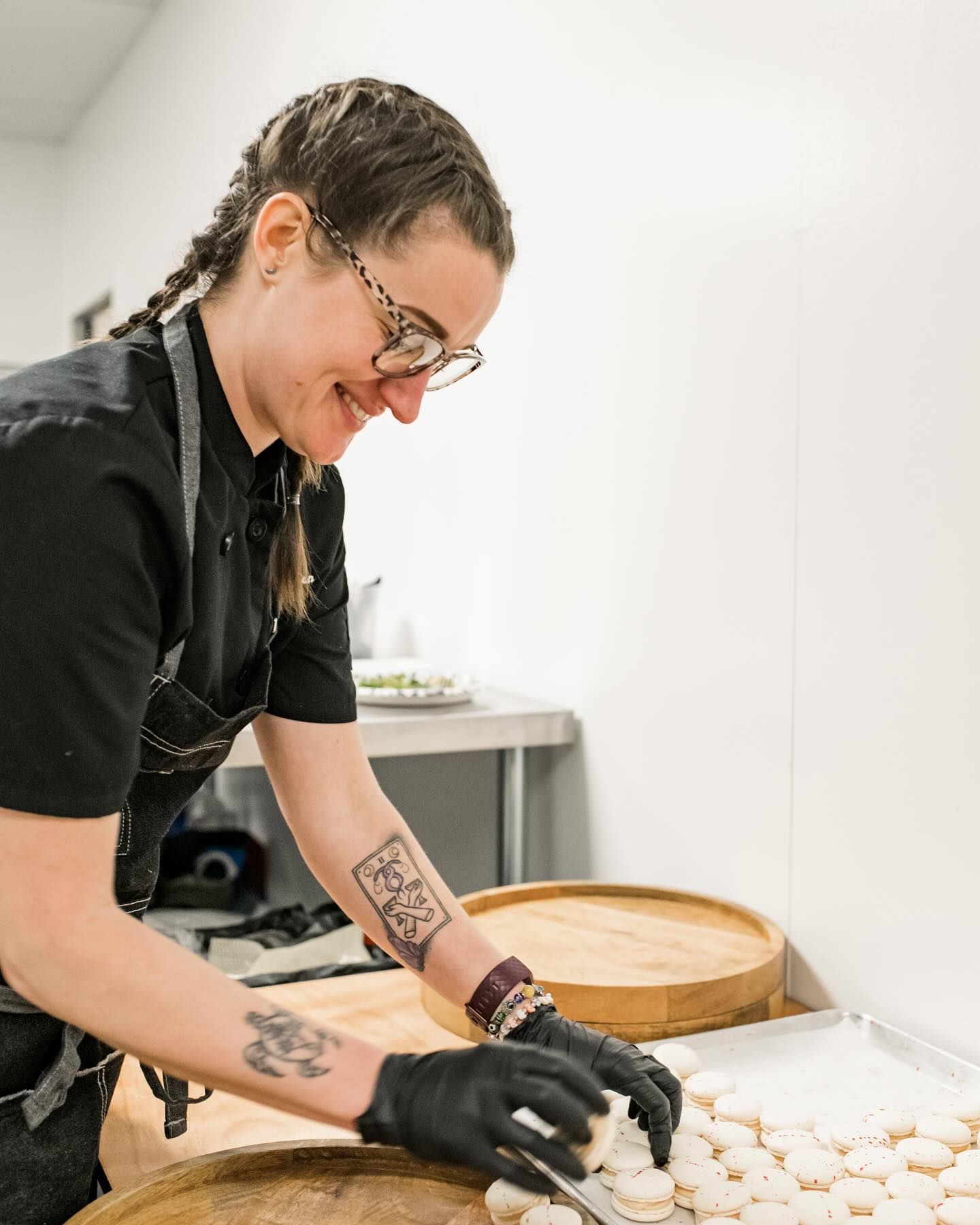 Chef placing pastries on a tray, wearing black gloves and uniform, with braided hair and a tattoo.