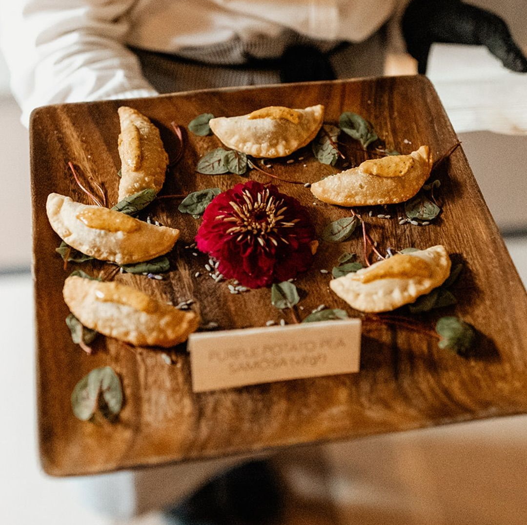 Wooden platter with purple potato samosas and a red flower.