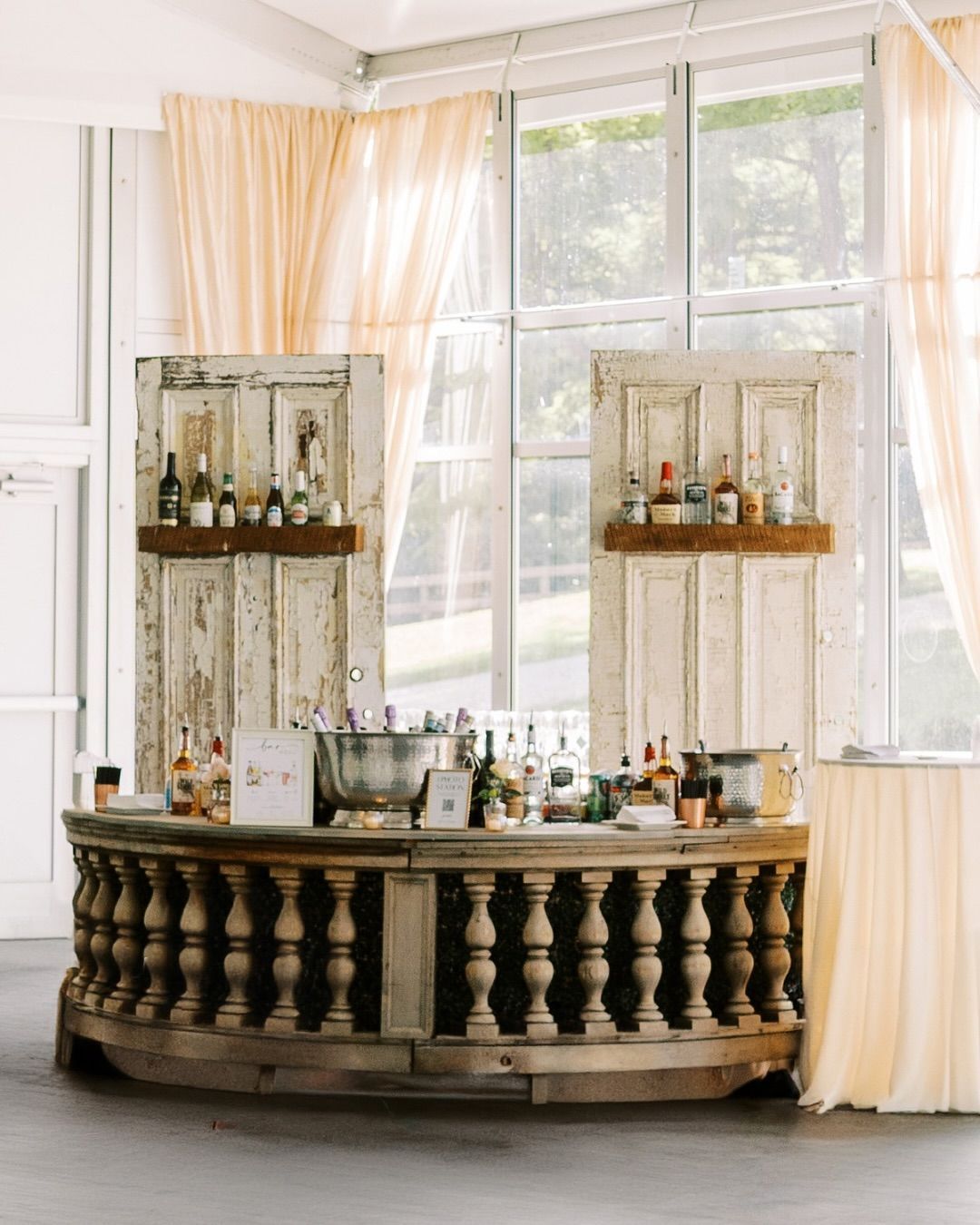 Rustic bar with bottles, set in front of large windows with draped curtains, and flanked by vintage door shelves for an Annapolis Catering Event