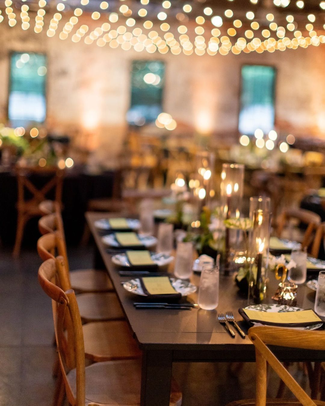 Long dinner table set for a gwith candles, lights, and wooden chairs in a brick-walled room.