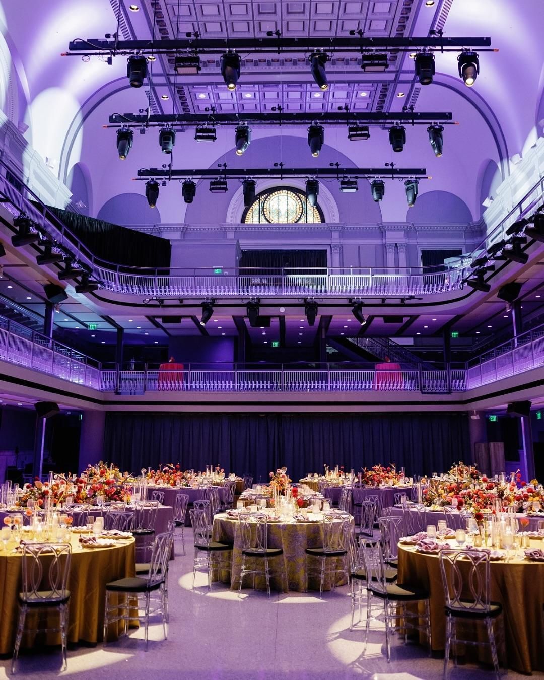 Formal event hall set with tables, gold tablecloths, and clear chairs under purple lighting.
