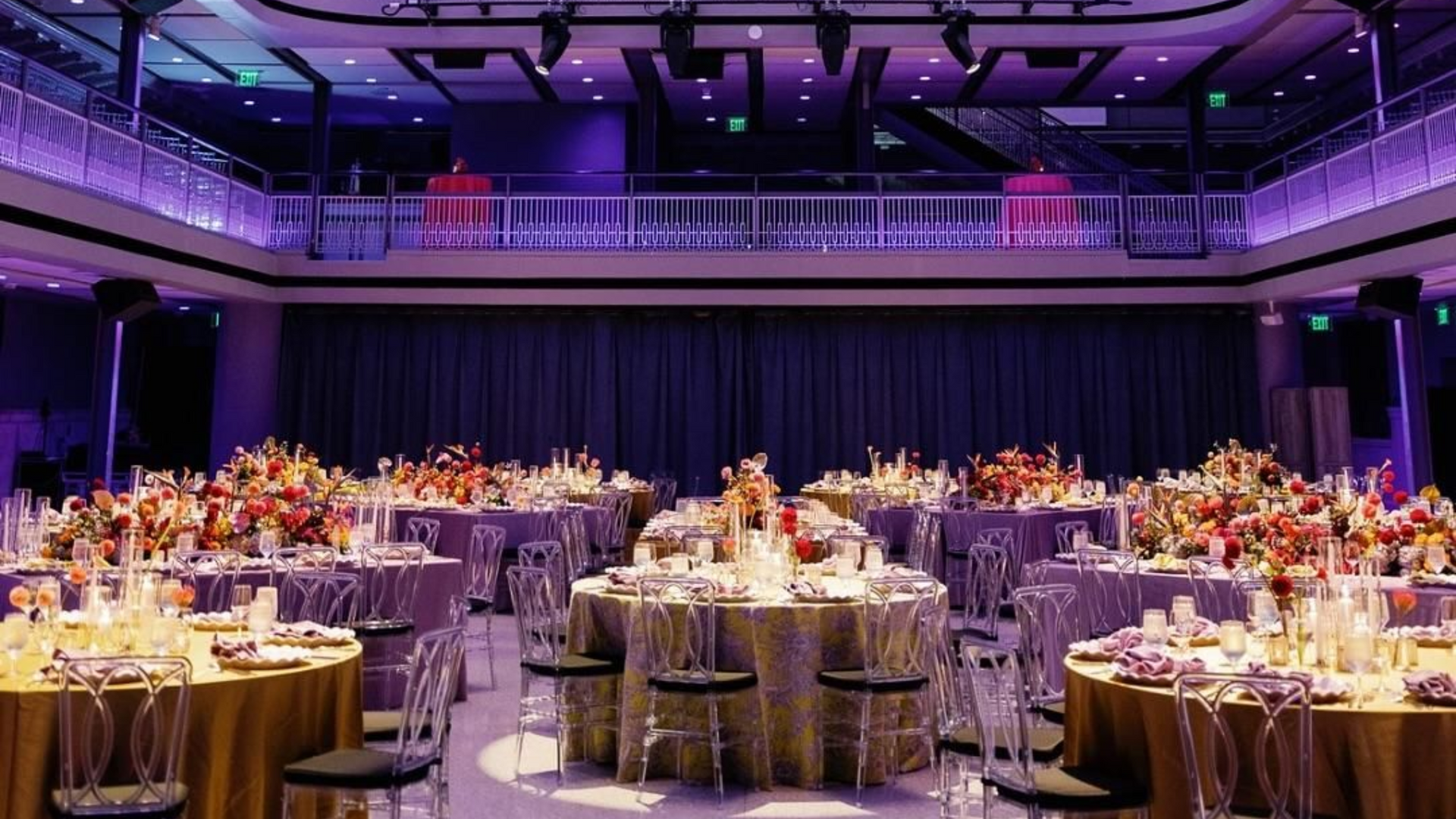 Elegant ballroom set for event, round tables with floral centerpieces, purple and gold decor.