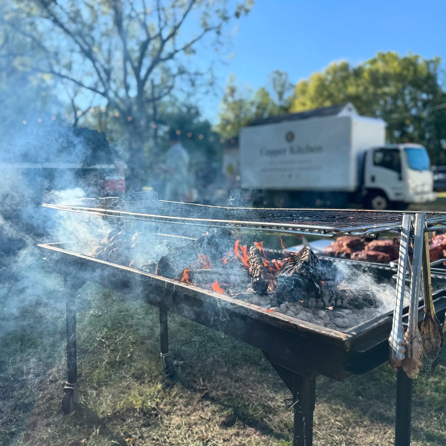 Large outdoor grill with burning wood, smoke, and food. A food truck is in the background.