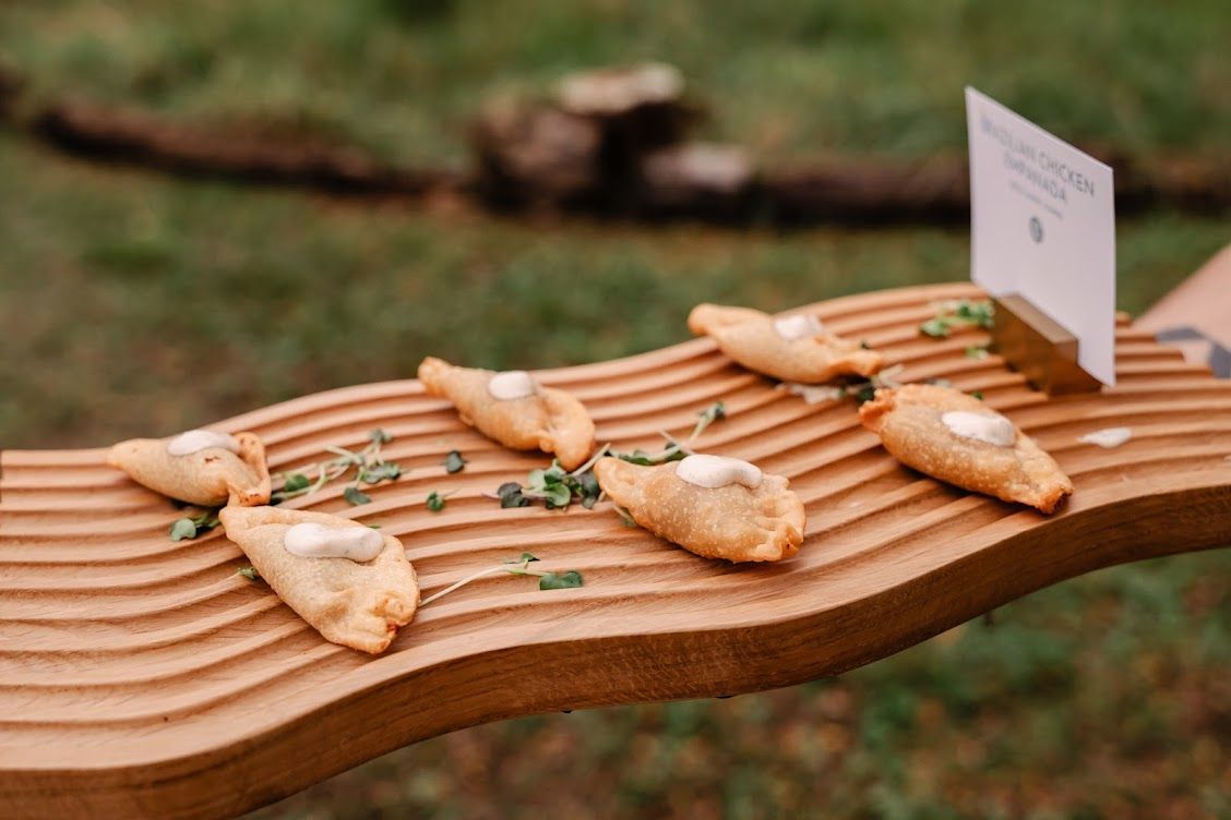 Appetizers on a wooden platter: six filled pastries with sauce, herbs, and a menu sign on grass.
