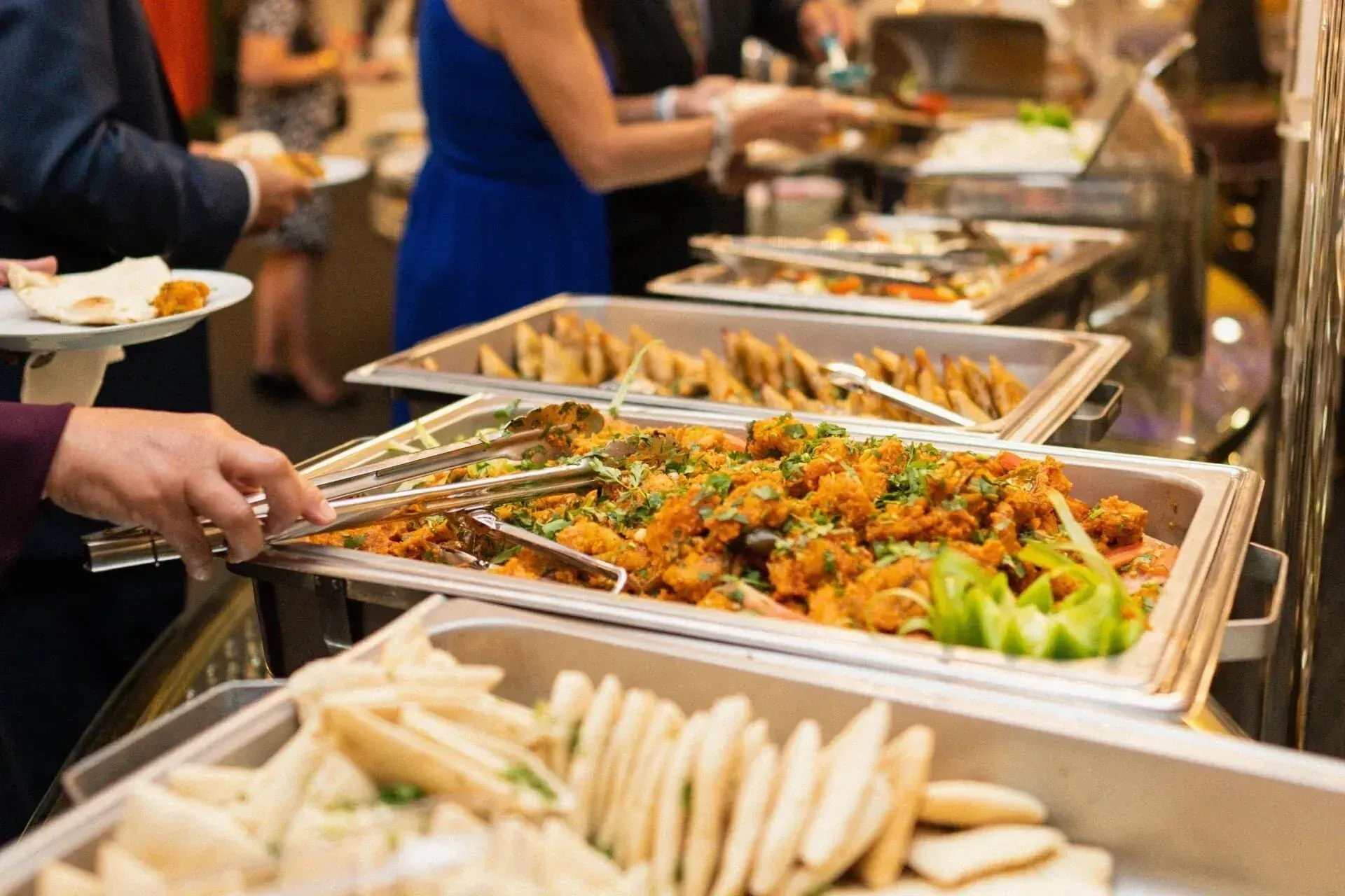 People at a buffet, selecting food with serving utensils. Trays of various dishes in view.