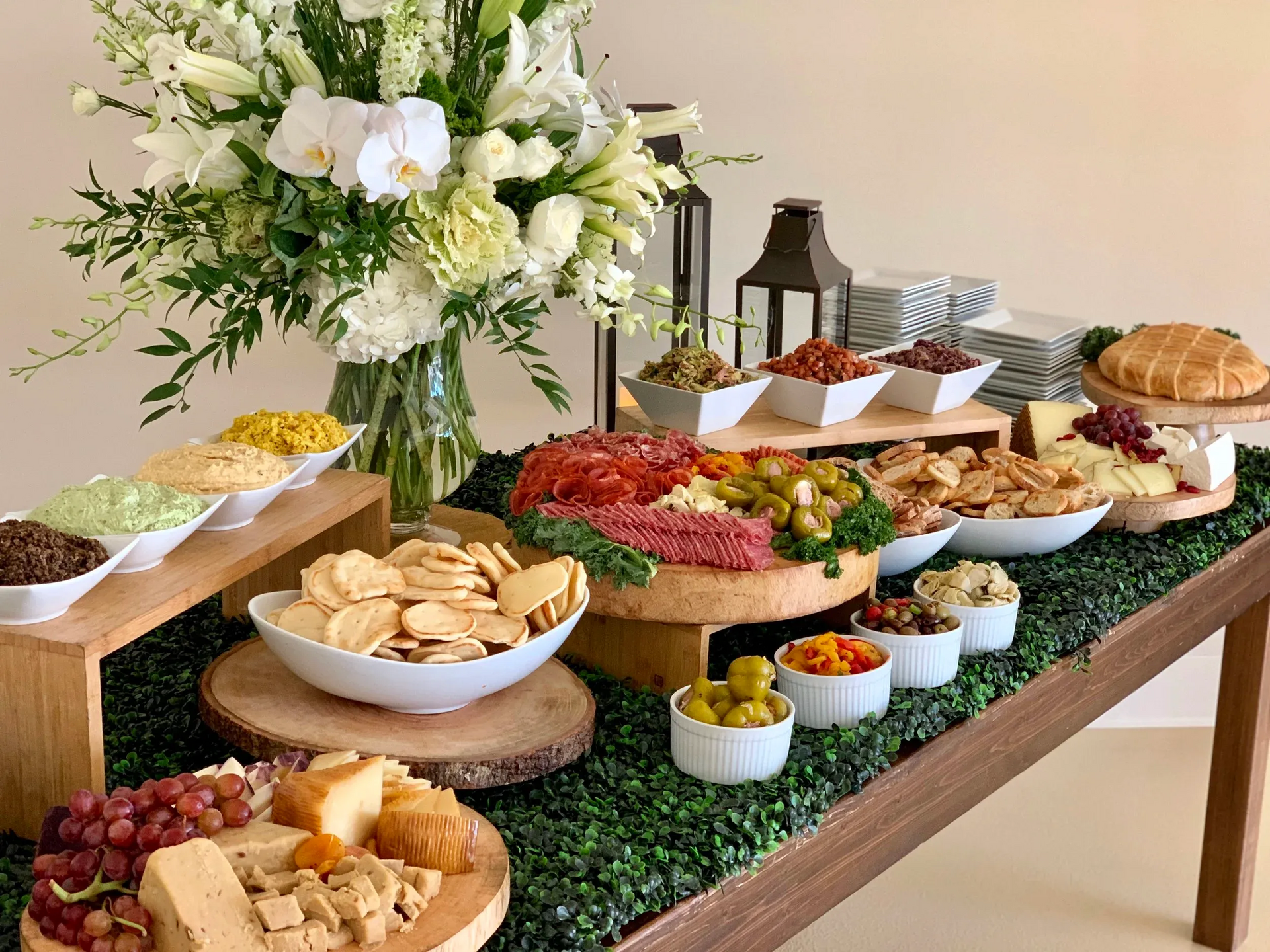 Charcuterie board on a table decorated with greenery and flowers. Contains dips, crackers, cheese, and olives.