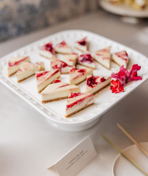 Triangular cheesecake bites with red swirls on a white platter, displayed with a flower and description card.
