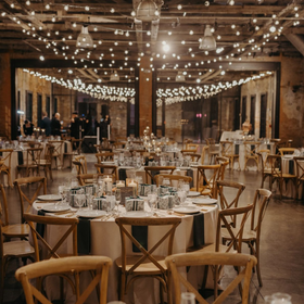 Wedding reception: round tables set with dishes, surrounded by cross-back chairs, under string lights in a rustic venue.