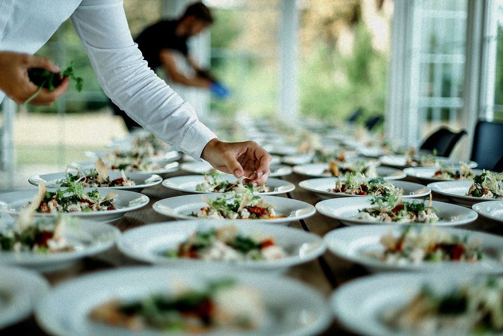 A chef in a white uniform garnishes plates of food arranged on a long table in a bright, outdoor setting.
