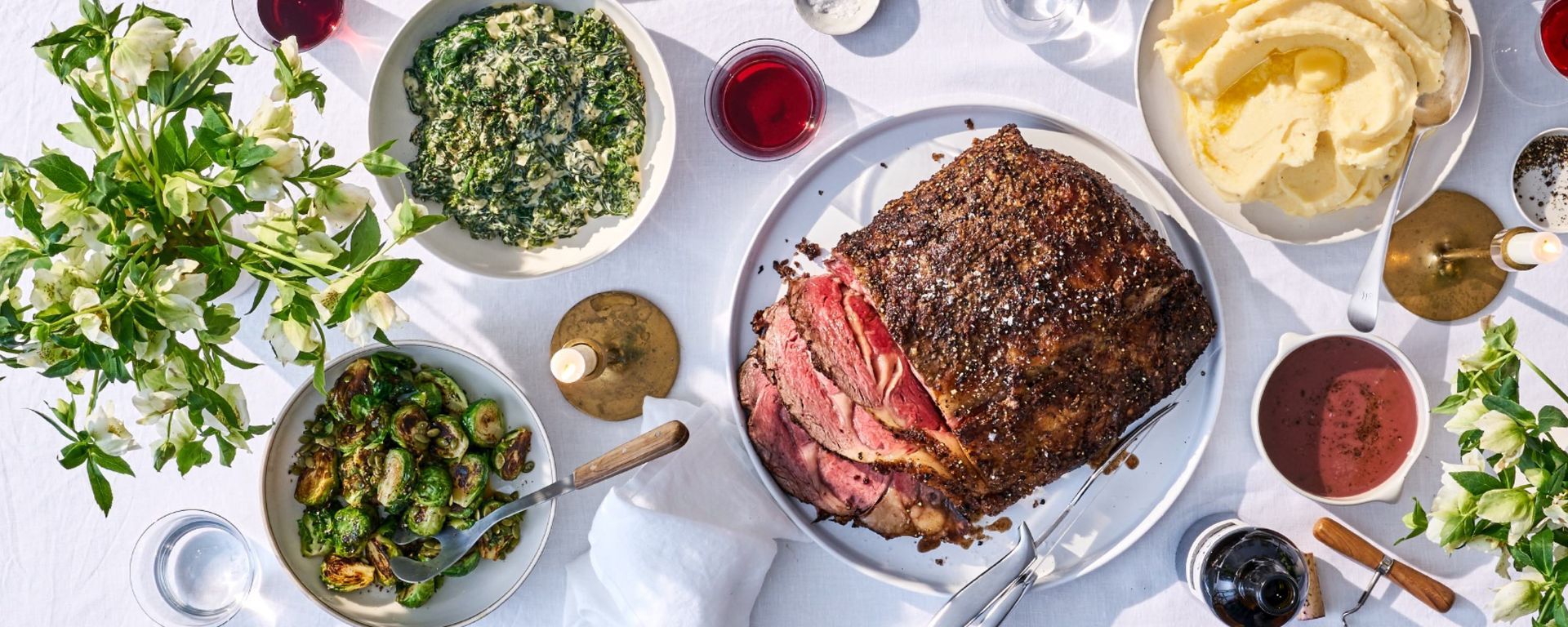 Overhead shot of a holiday meal spread on a white tablecloth, including sliced roasted meat, mashed potatoes, and side dishes.