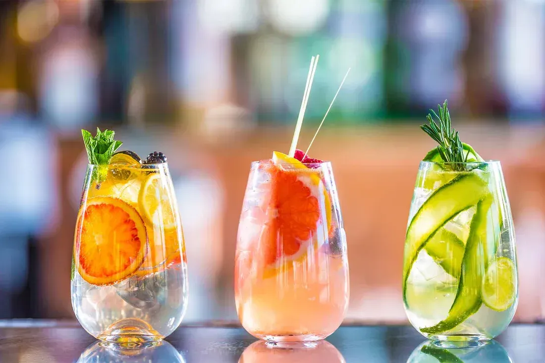 Three fruit and herb infused cocktails on a bar, with colorful fruit slices and garnishes.
