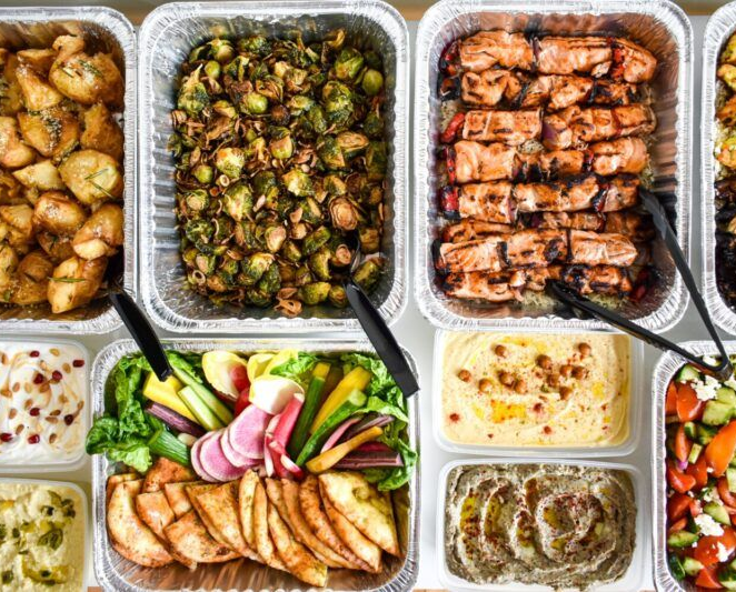 Prepared food in aluminum trays: potatoes, Brussels sprouts, chicken, hummus, and vegetables.