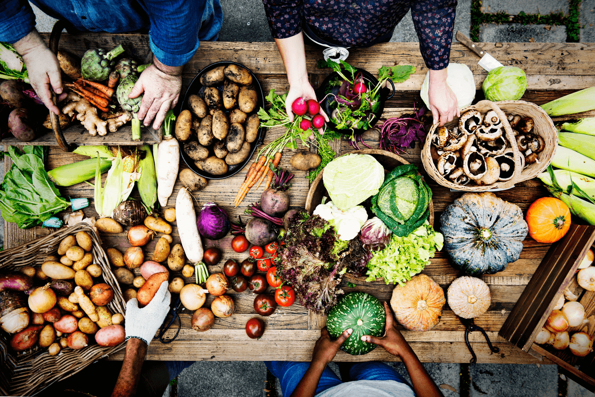 People arranging various fresh vegetables, potatoes, and mushrooms on a large rustic wooden table.