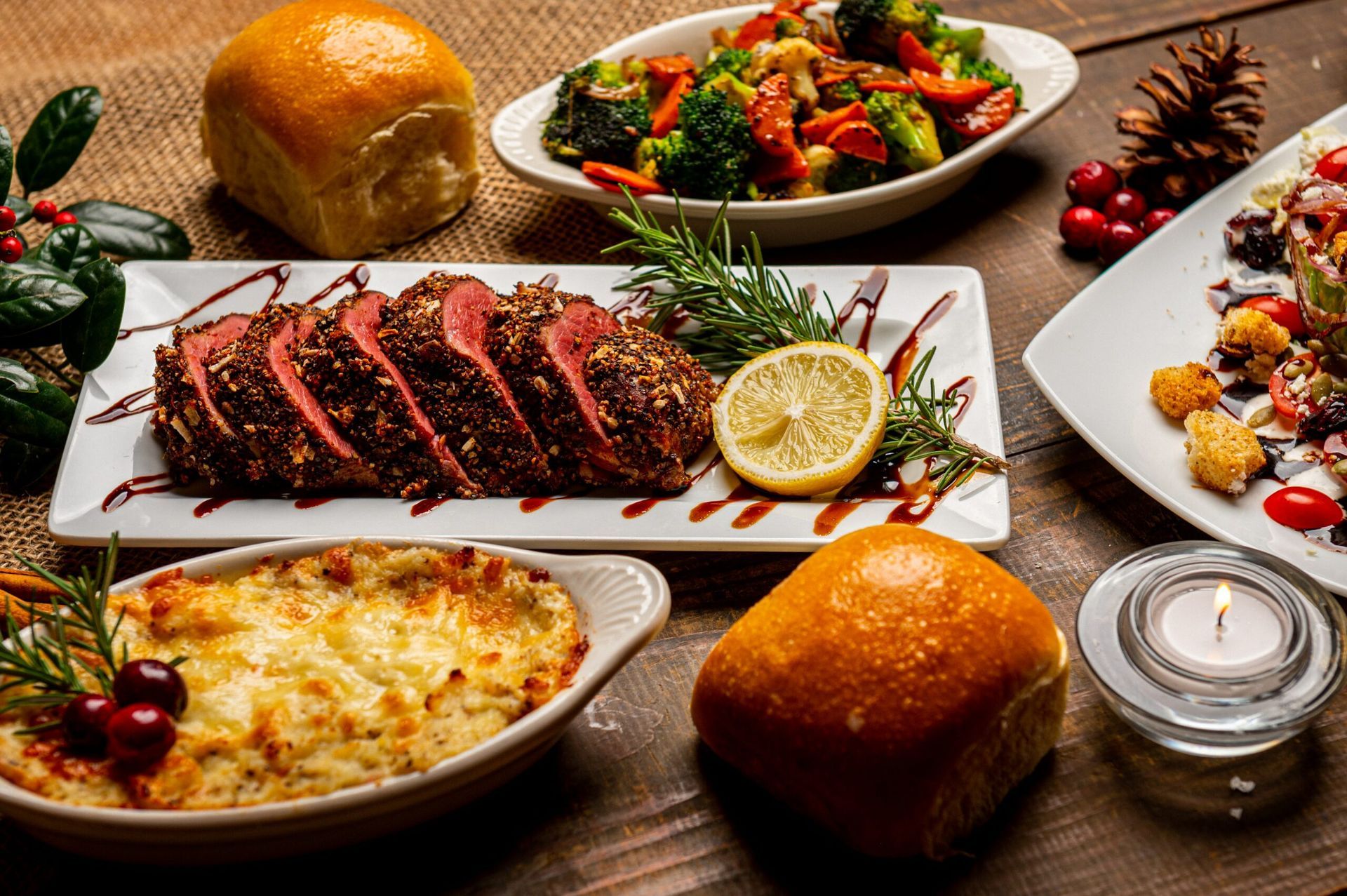Holiday meal with sliced roast, vegetables, rolls, and a side dish, on a wooden table.