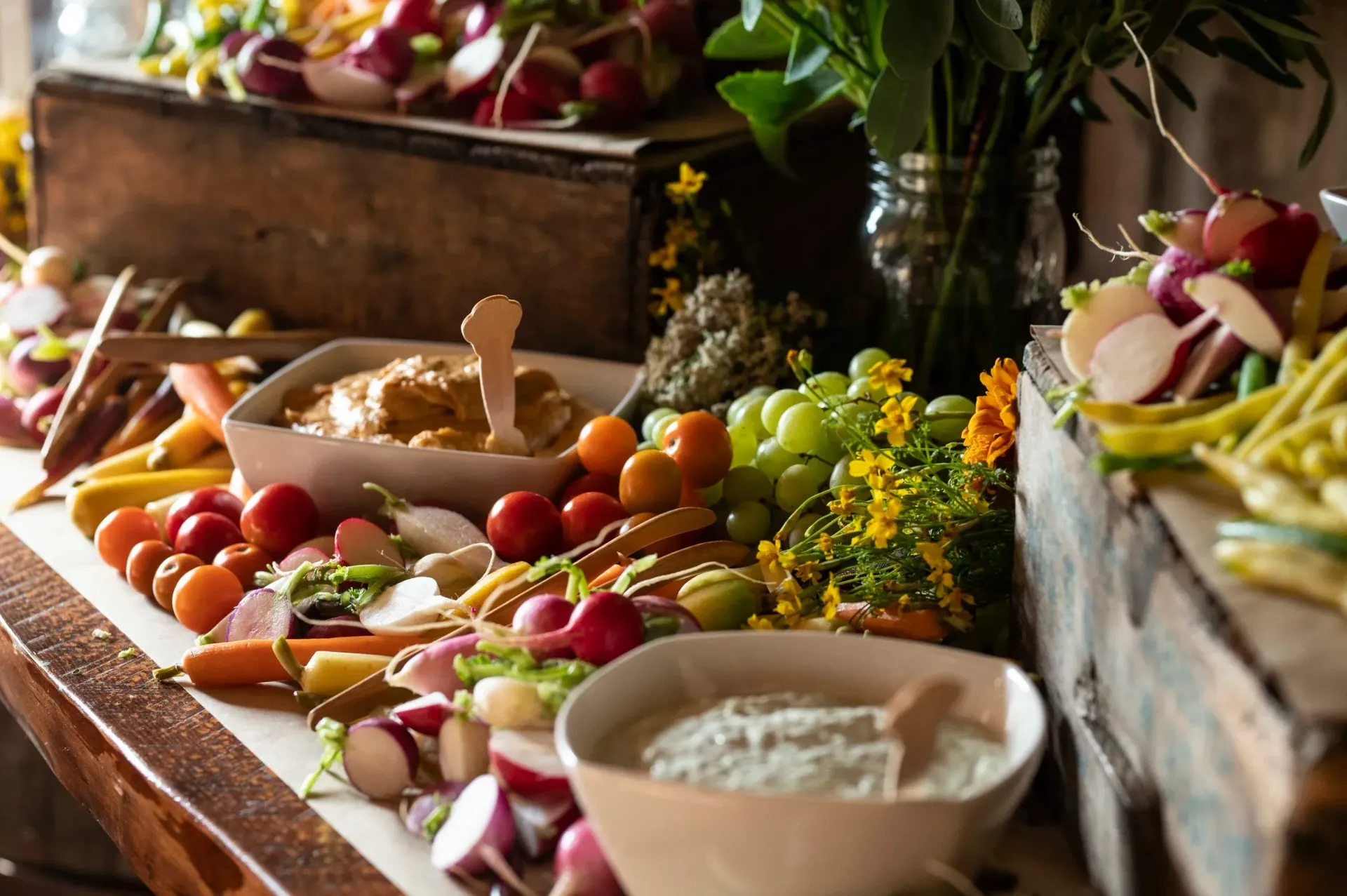 A rustic wooden table displays a colorful spread of fresh vegetables, grapes, and bowls of dip for a gathering.
