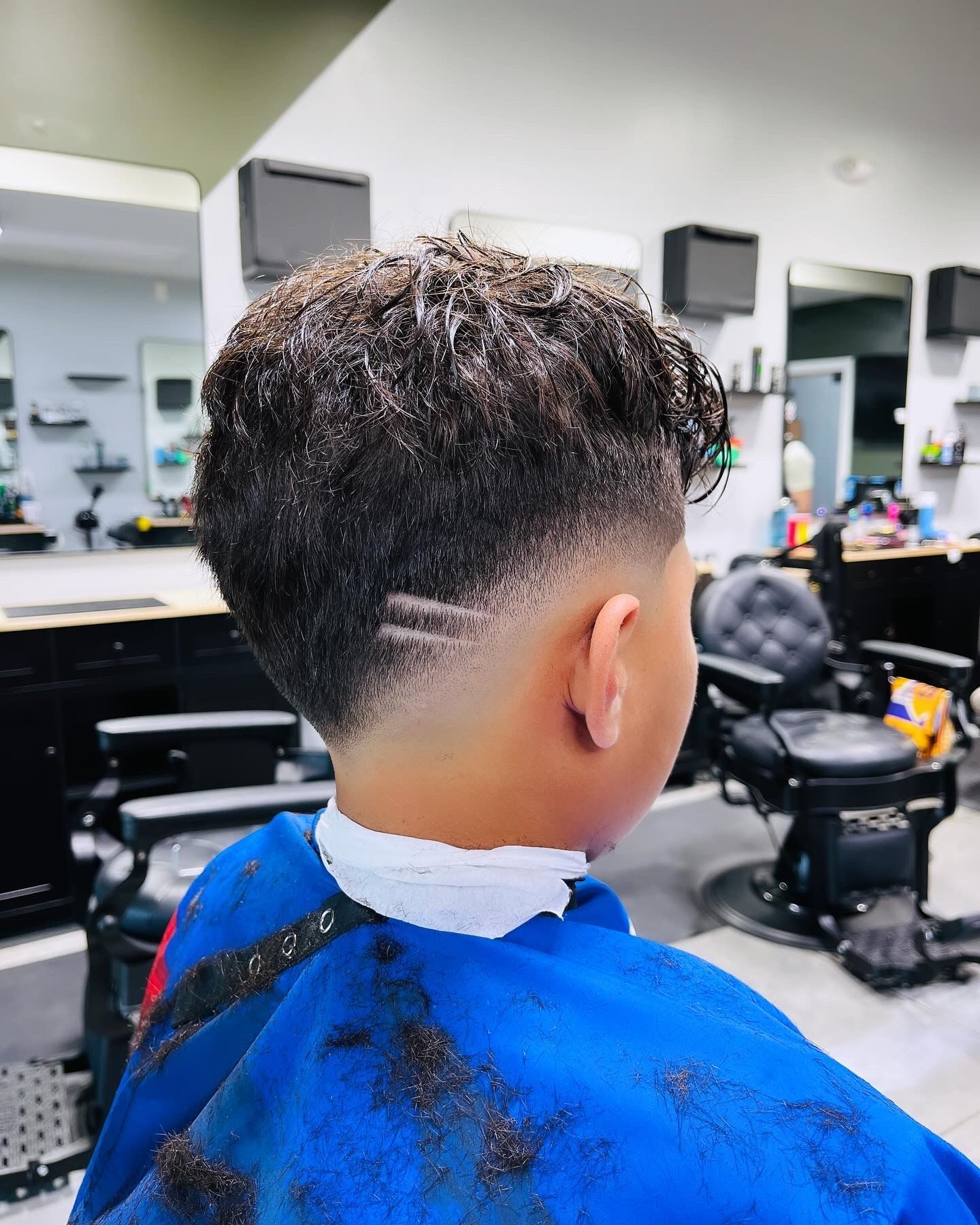 Boy with a faded haircut, designs on the side, inside a barber shop, wearing a blue cape.