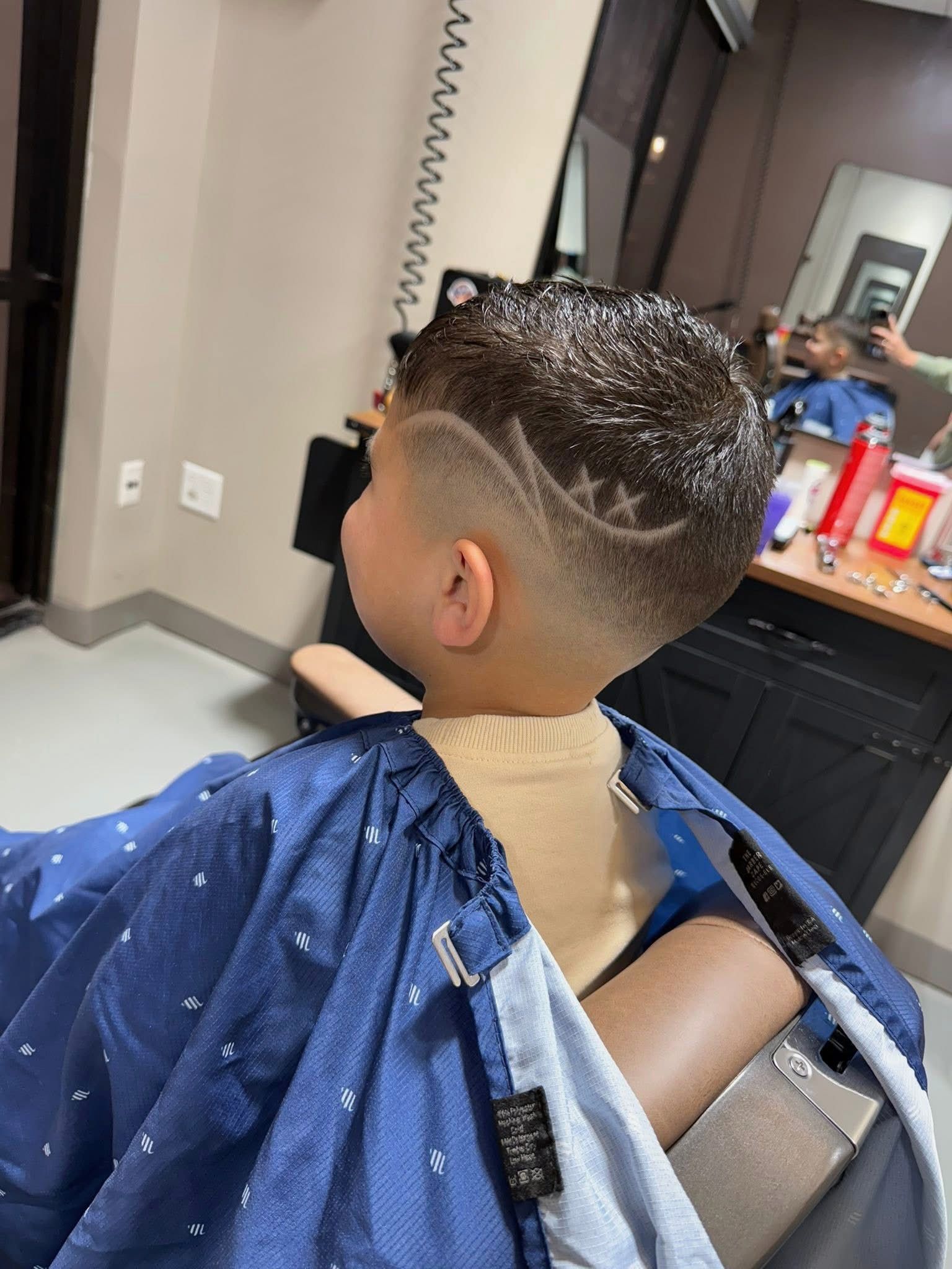 Boy with a star design haircut, wearing a blue cape, in a barber shop.