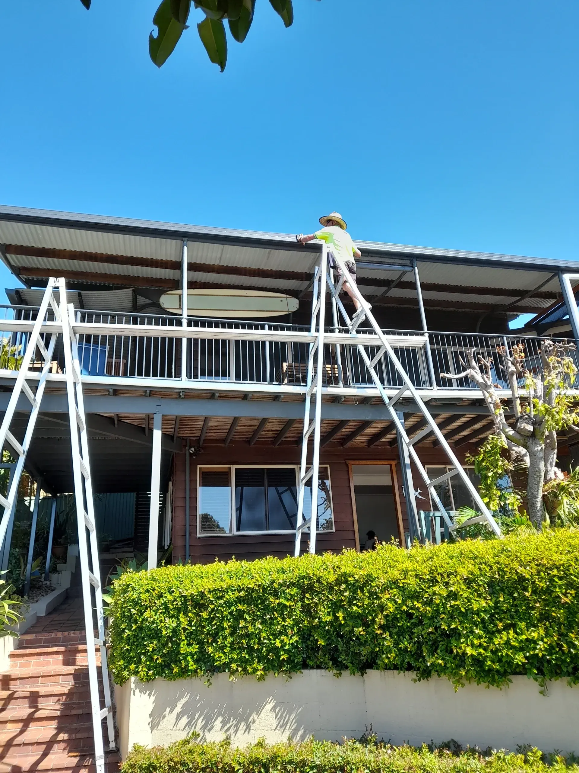 Person on ladder repairing roof of two-story house with a balcony, blue sky in the background.