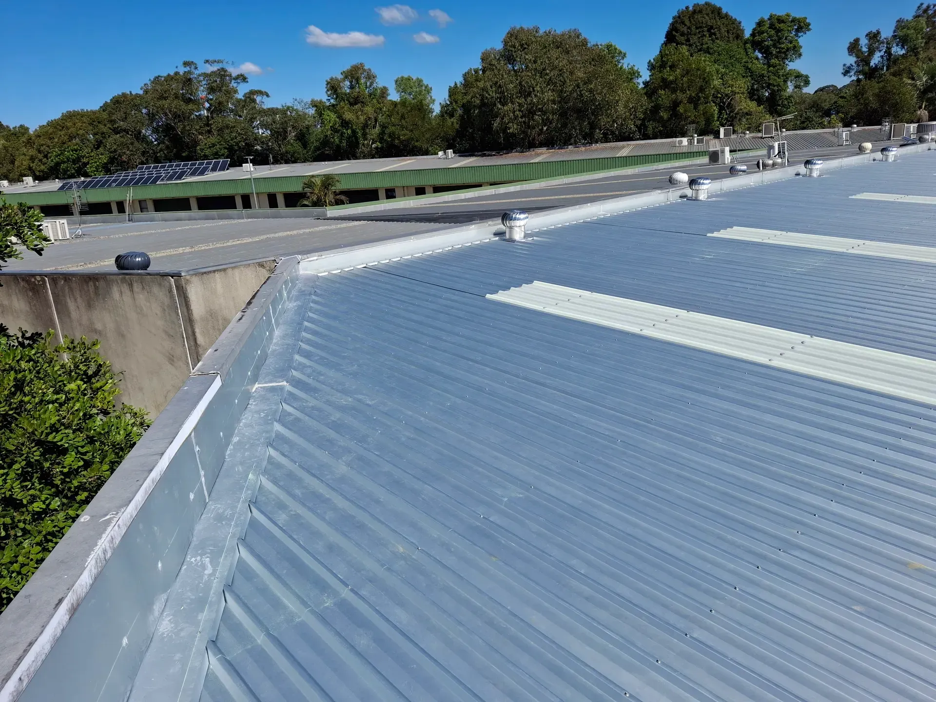 Metal roof with a corrugated design, a blue-gray color, and white accent stripes. Trees and a concrete building in the background.