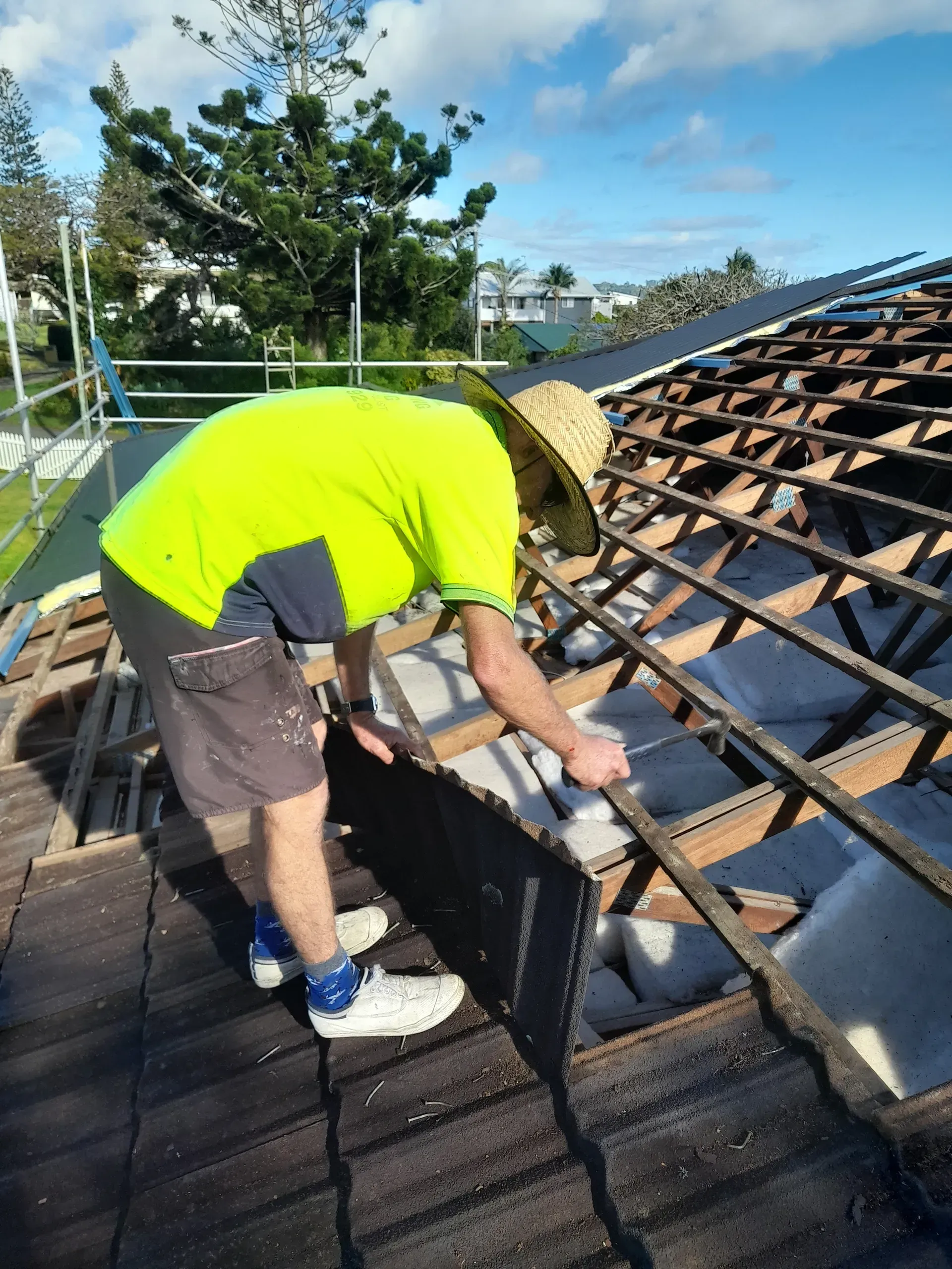 Person on a rooftop in a straw hat, replacing a roof tile. He is wearing a neon shirt and shorts.