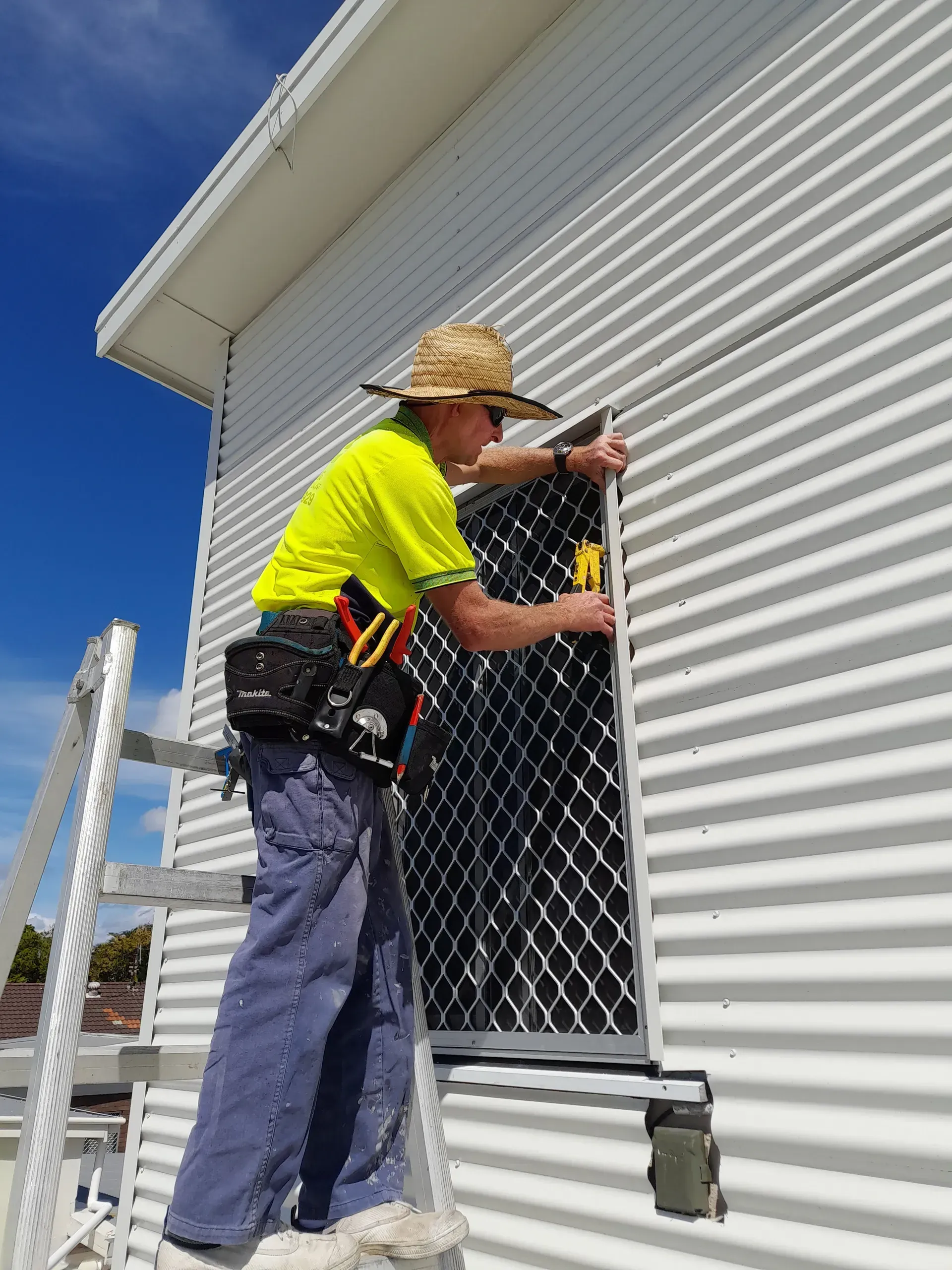 A person in a straw hat installs a security door on a white corrugated building, standing on a ladder.