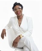 Woman in white suit, seated, smiling, wearing glasses and jewelry.
