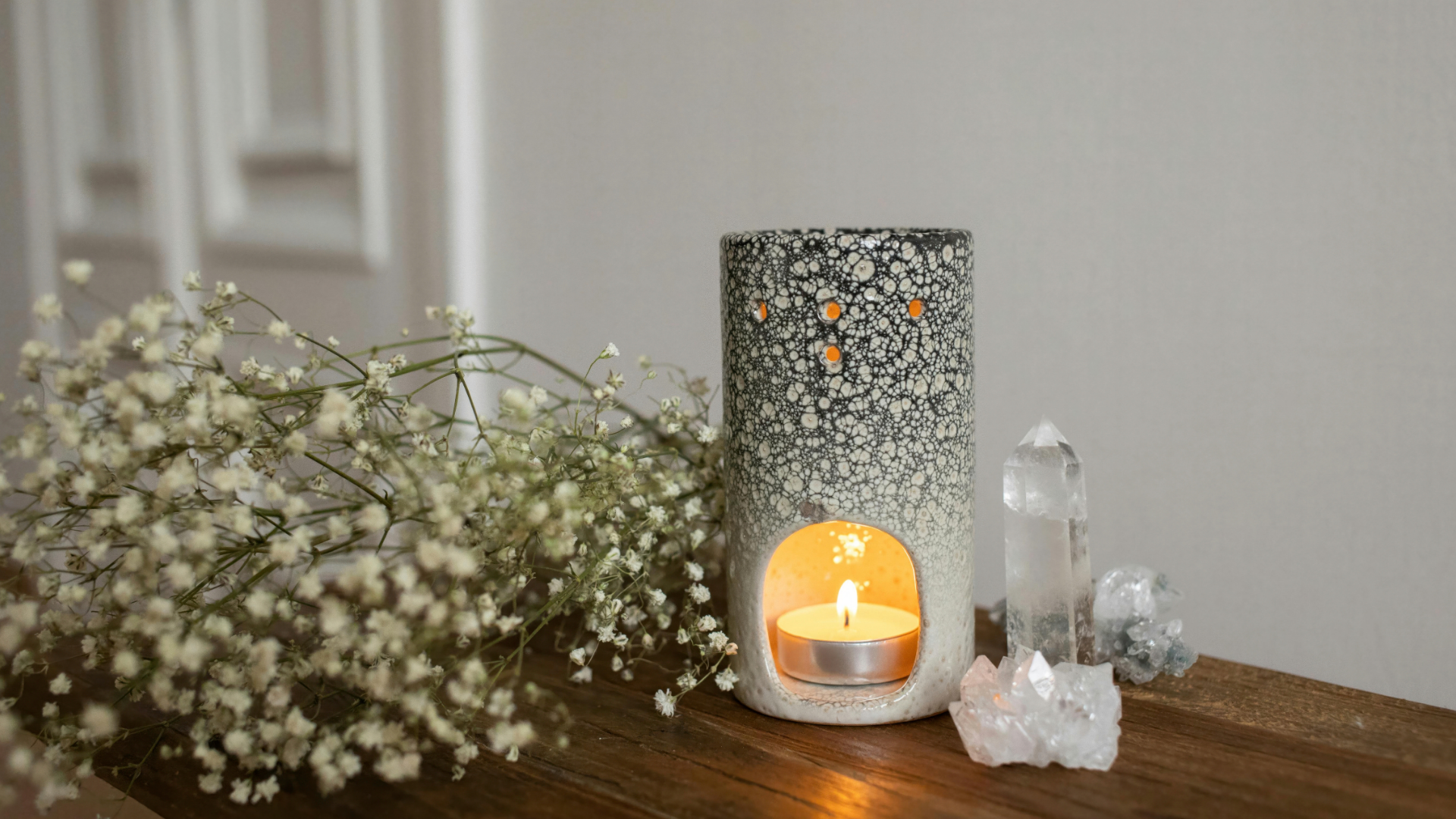 Ceramic oil warmer with a lit candle, crystals, and flowers on a wooden surface.