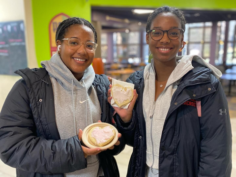Two smiling people wearing hoodies and jackets hold small, heart-shaped frosted cookies.