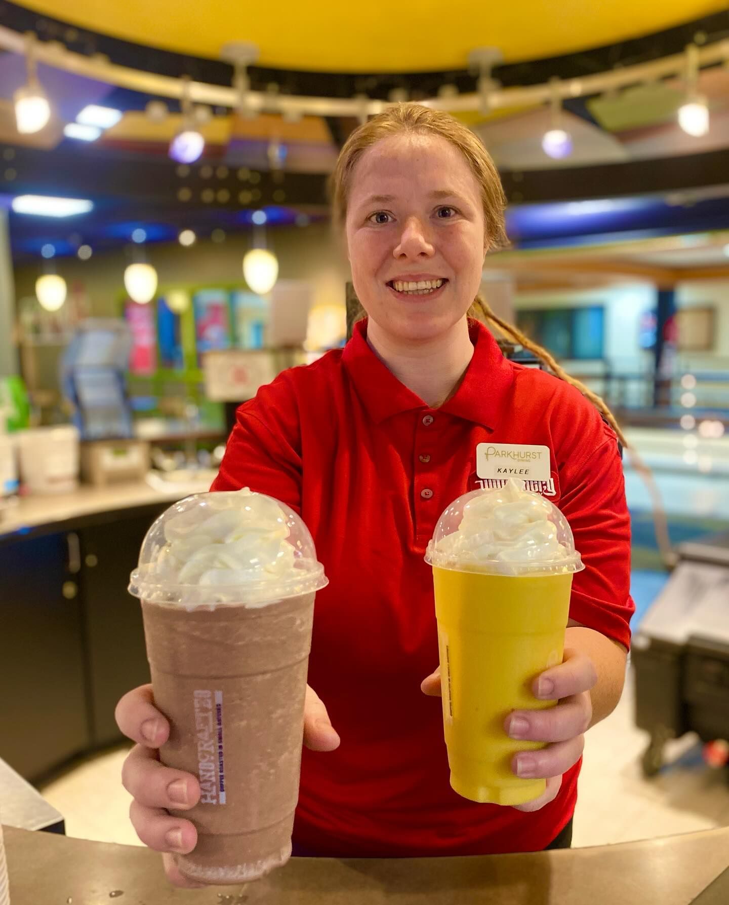 Woman in red shirt holding two whipped cream-topped drinks in a brightly lit food service area.