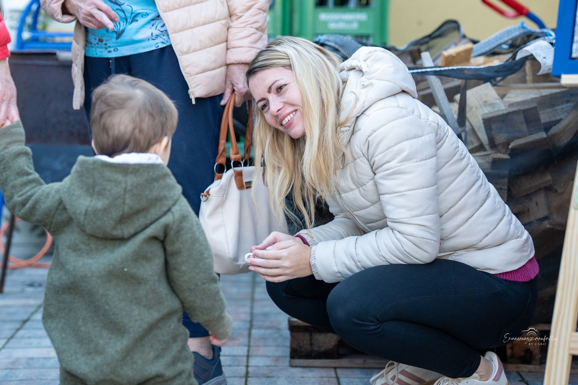 Frau hockt im Freien und lächelt ein Kleinkind an, das sie an der Hand hält.