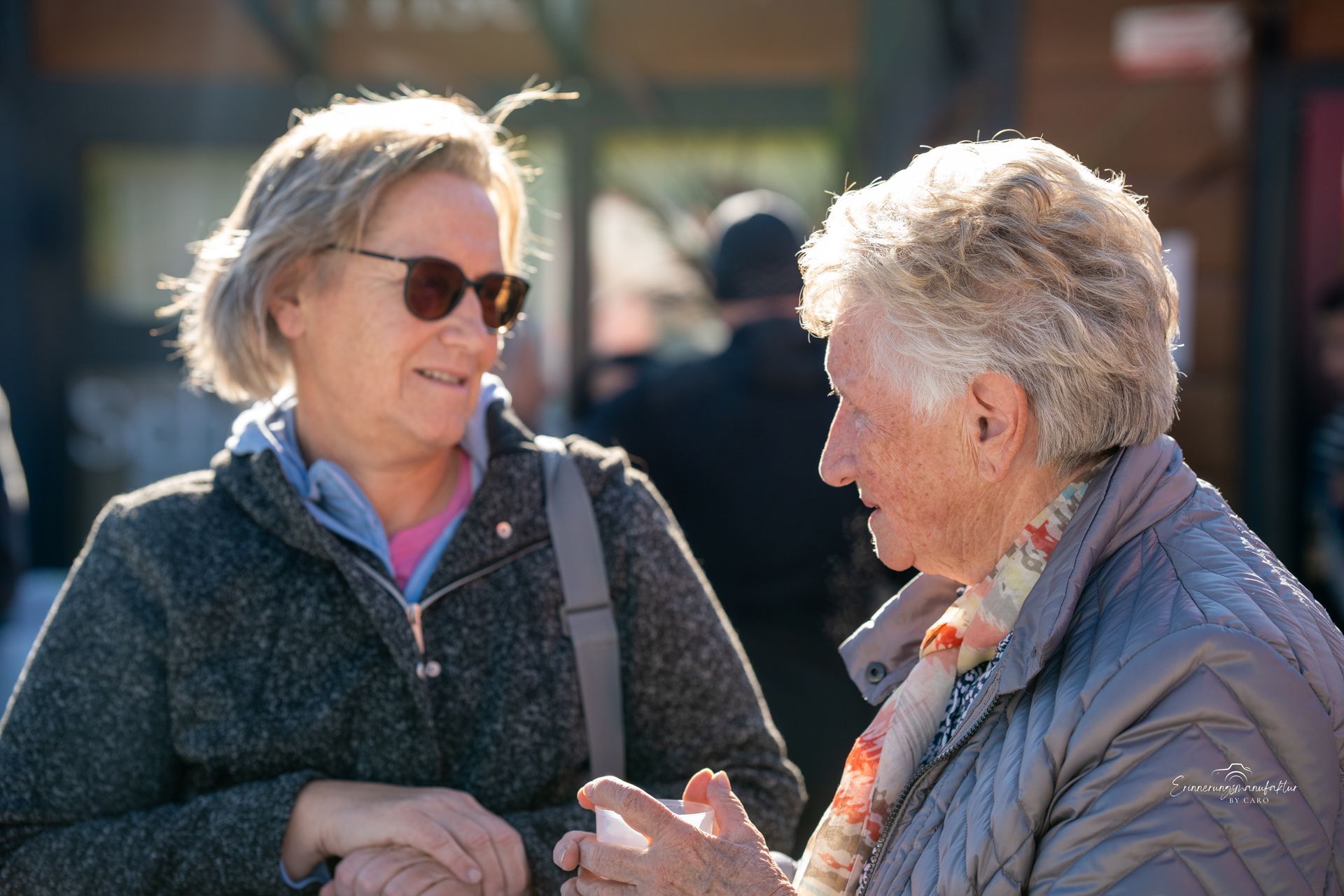 Zwei Frauen unterhalten sich im Freien; die eine trägt eine Sonnenbrille und einen Mantel