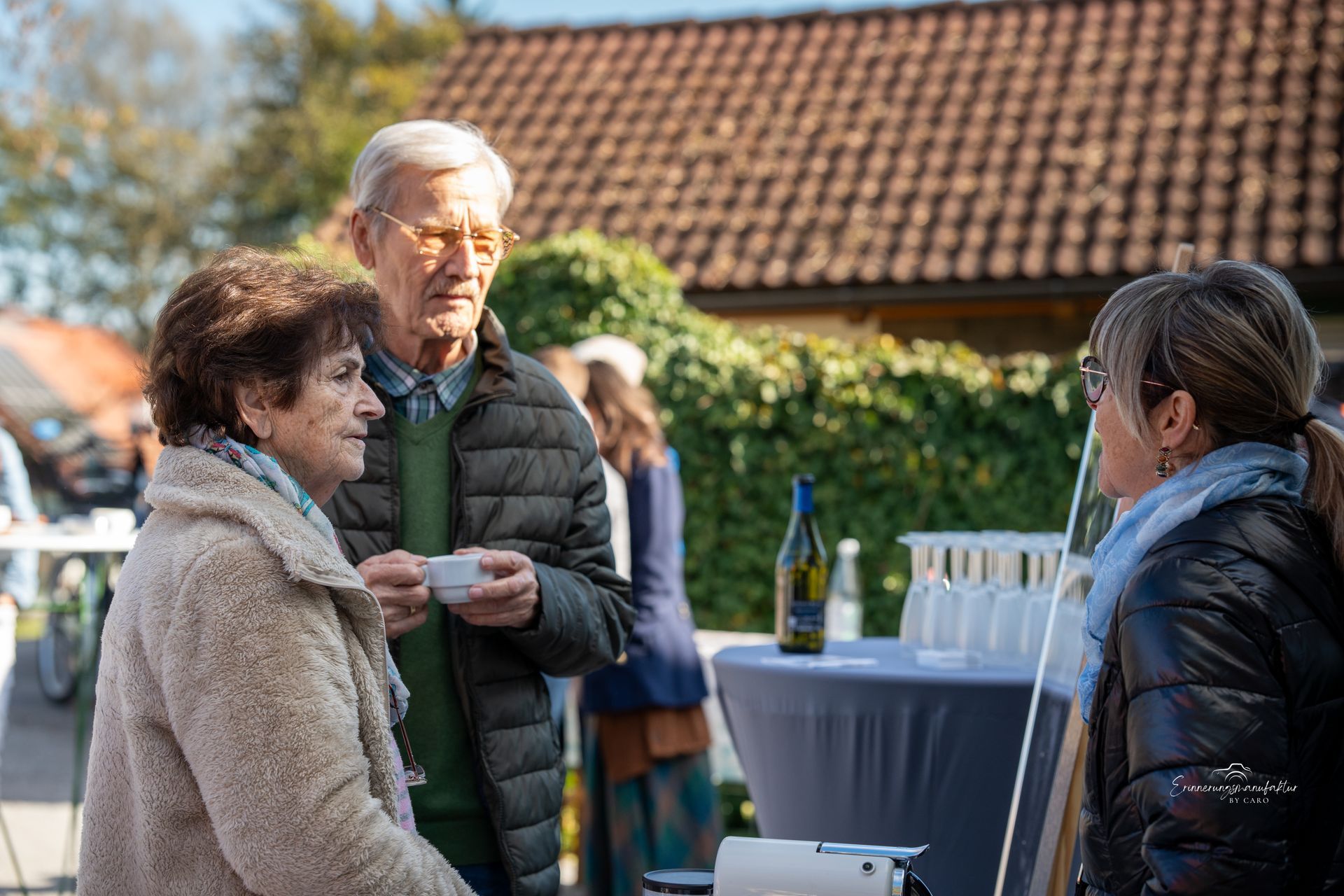 Drei Personen unterhalten sich im Freien an einem Tisch mit Getränken; ein Mann hält eine Tasse.