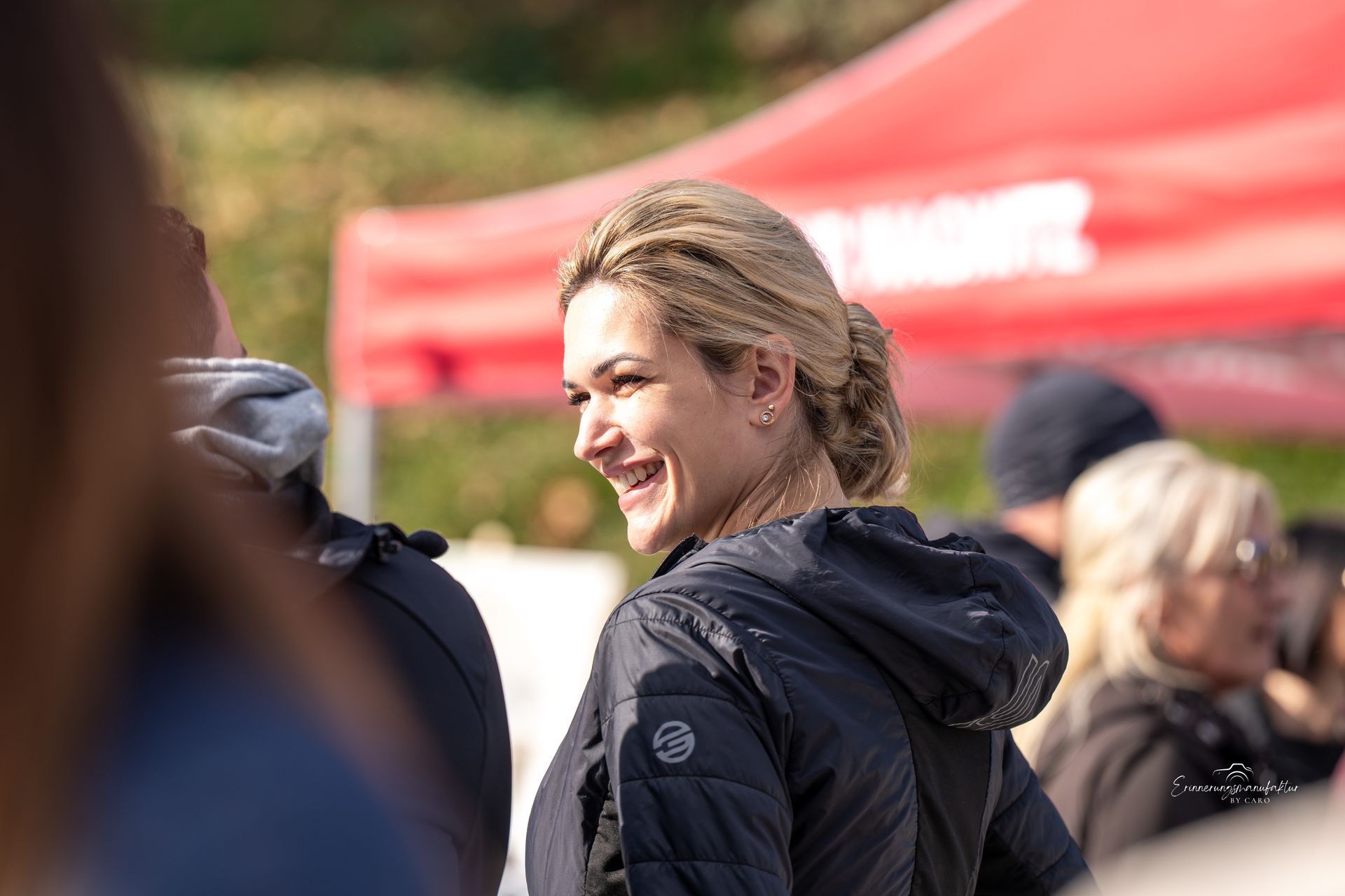 Frau mit blonden Haaren lächelt, trägt eine schwarze Jacke, im Freien.