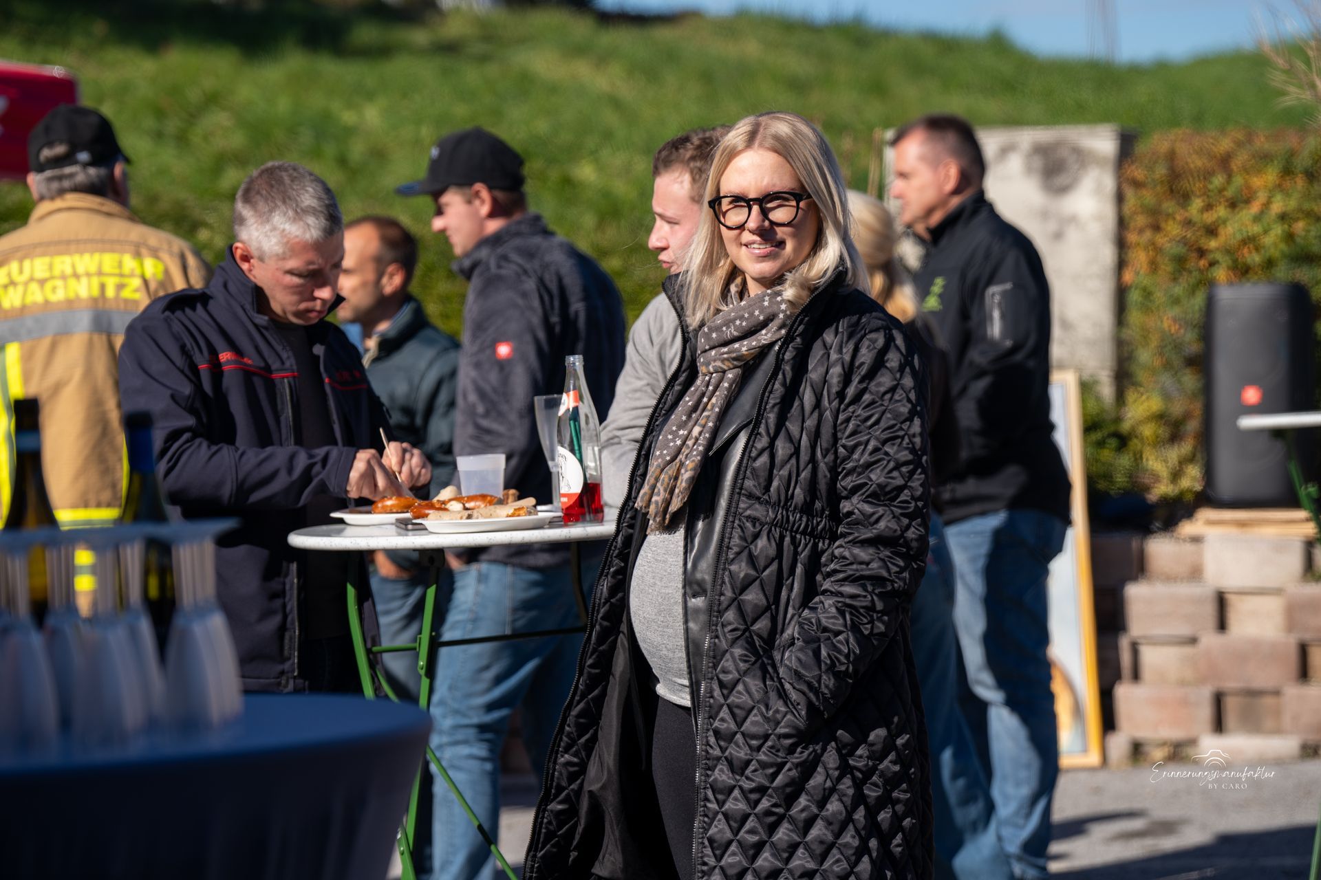 Eine Frau mit Brille und Steppmantel lächelt bei einer Zusammenkunft im Freien.