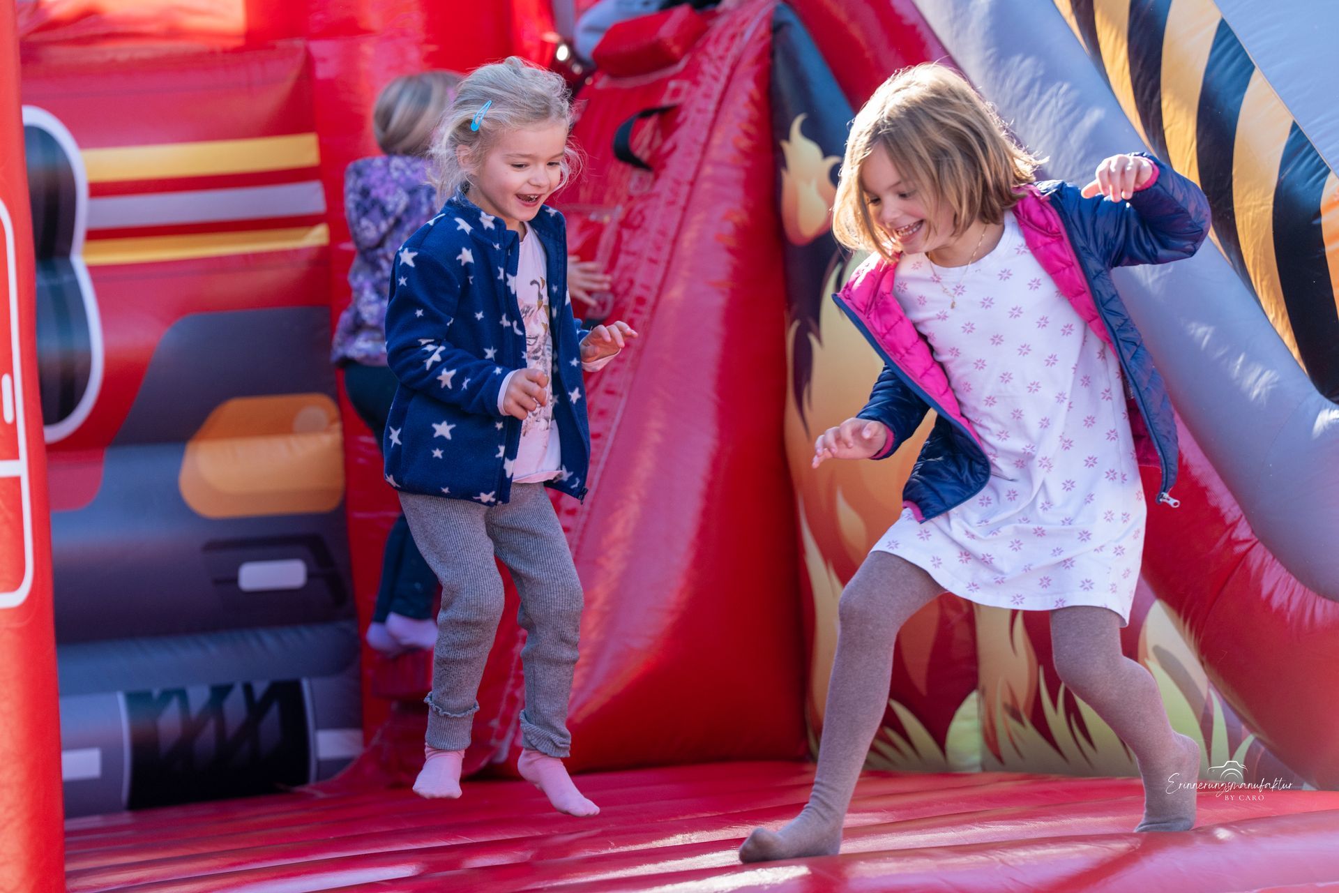 Zwei Mädchen springen und lachen auf einer roten Hüpfburg.