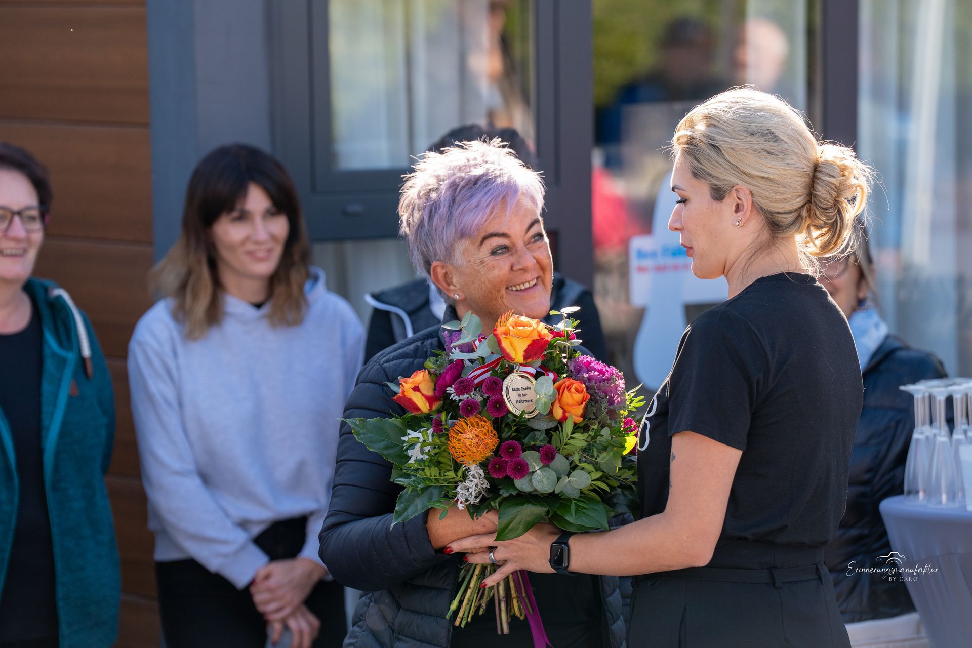 Eine Frau mit lila Haaren lächelt und erhält vor einem Gebäude Blumen von einer anderen Frau.