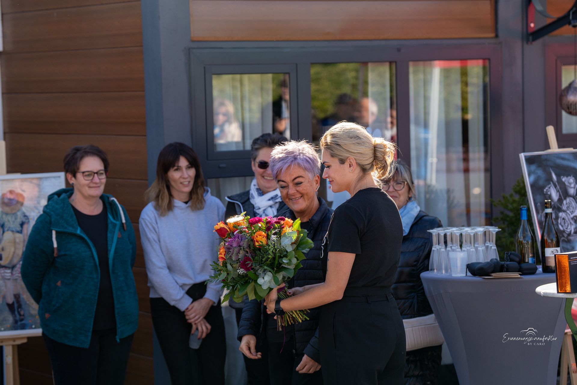 Eine Frau erhält von einer anderen Frau Blumen vor einem Gebäude, umgeben von Schaulustigen.