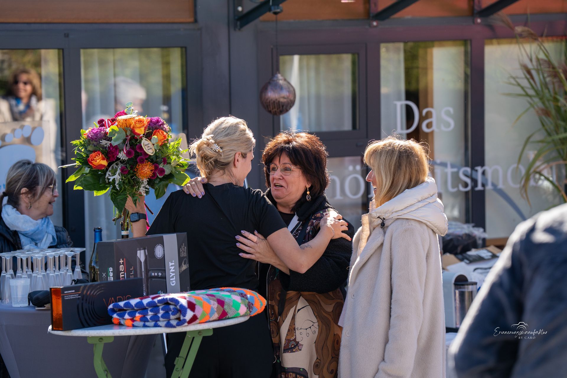 Drei Frauen umarmen sich, eine von ihnen hält Blumen, vor einem Gebäude mit der Aufschrift „Das“.