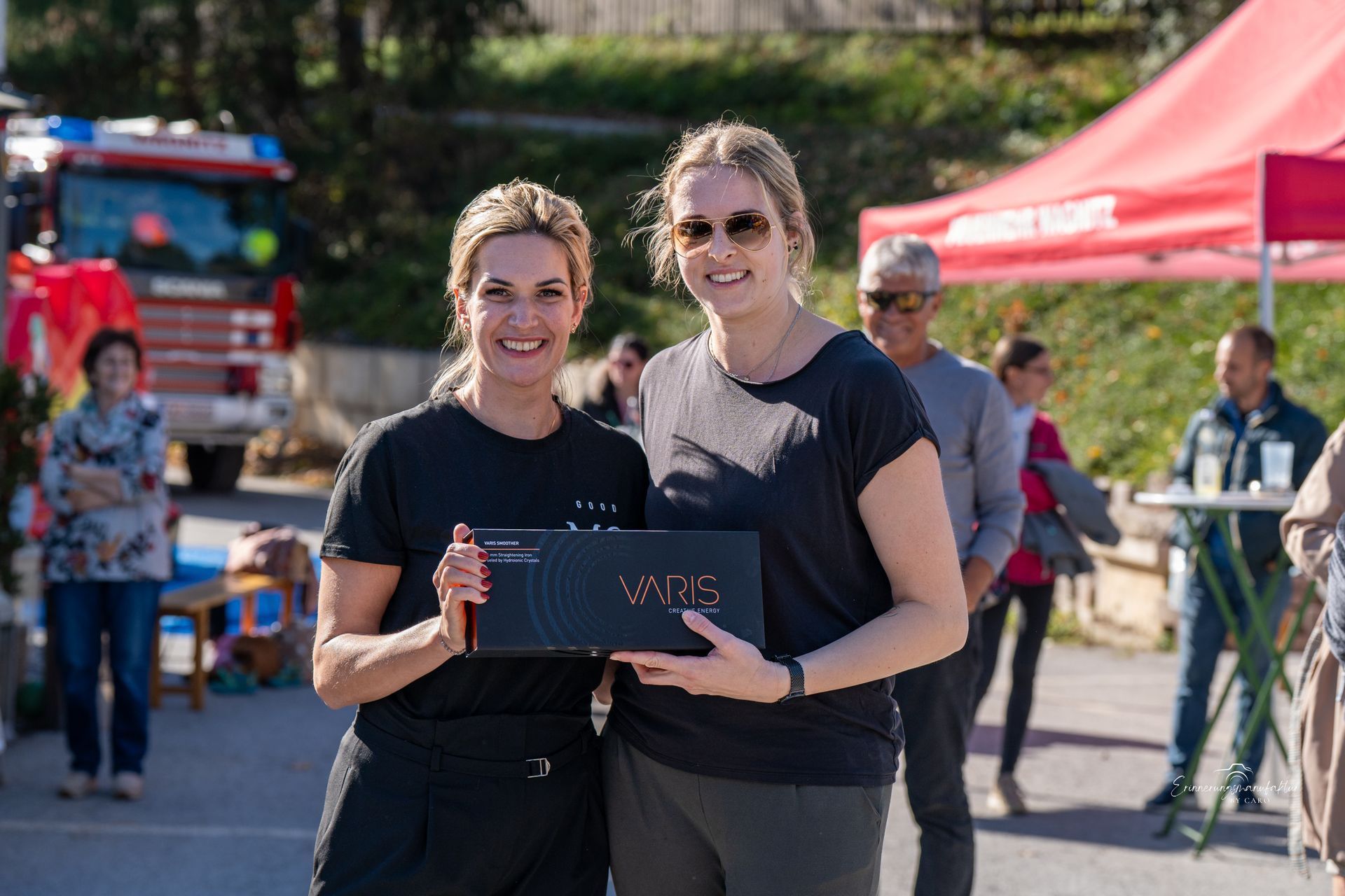 Zwei Frauen halten eine Geschenkbox und lächeln im Freien neben einem Feuerwehrauto und einem roten Zelt.