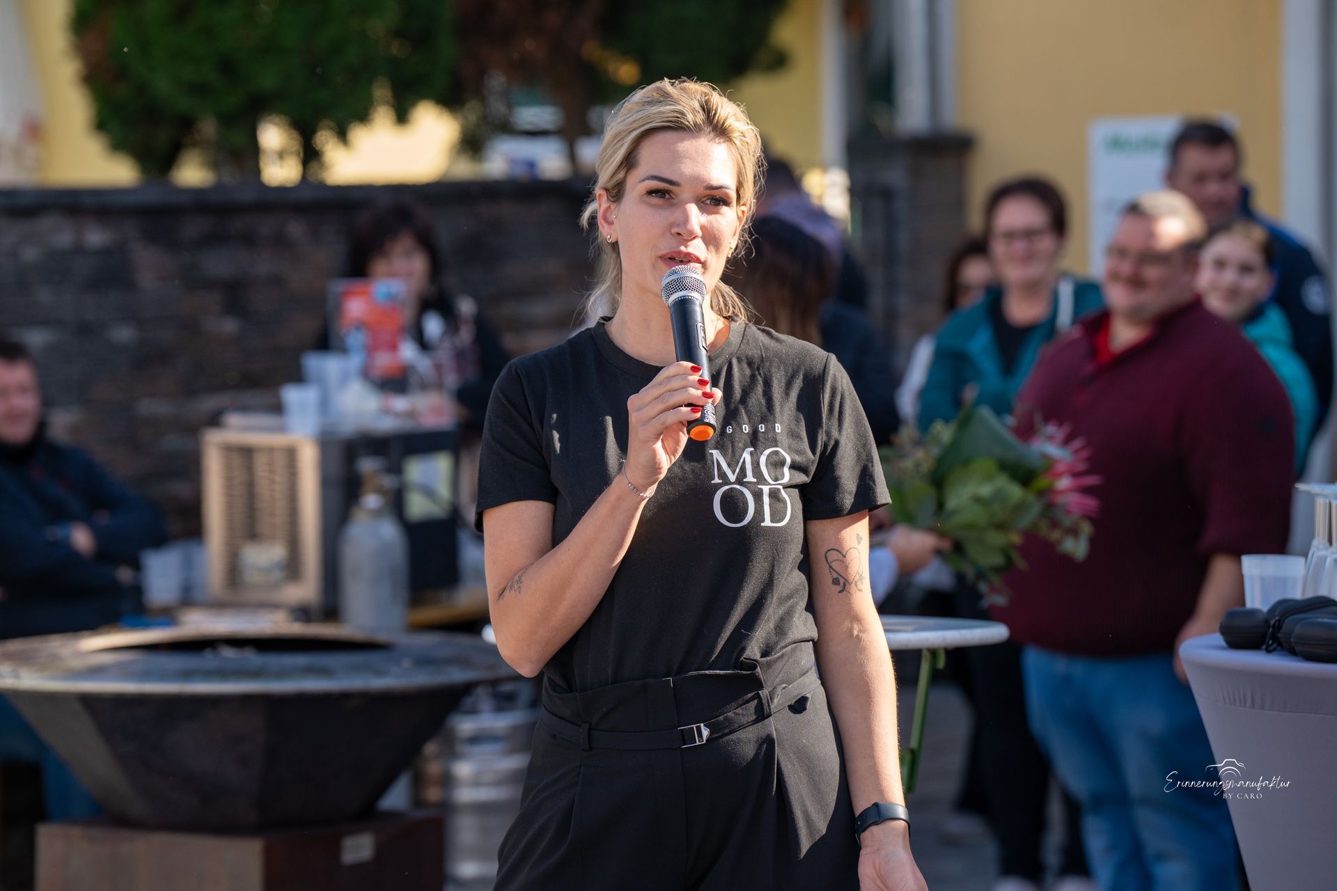 Frau spricht im Freien in ein Mikrofon; Personen im Hintergrund.