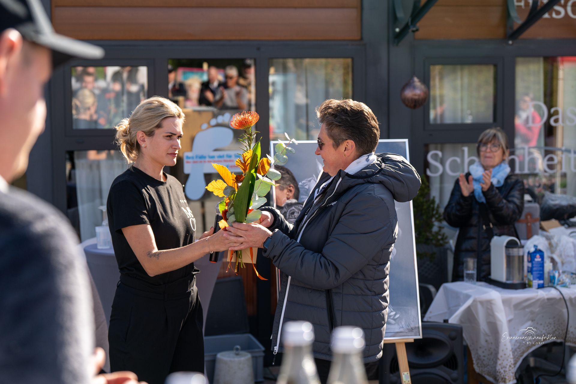 Eine Frau in schwarzem Kleid überreicht im Freien einer anderen Frau Blumen. Umstehende beobachten das Geschehen.