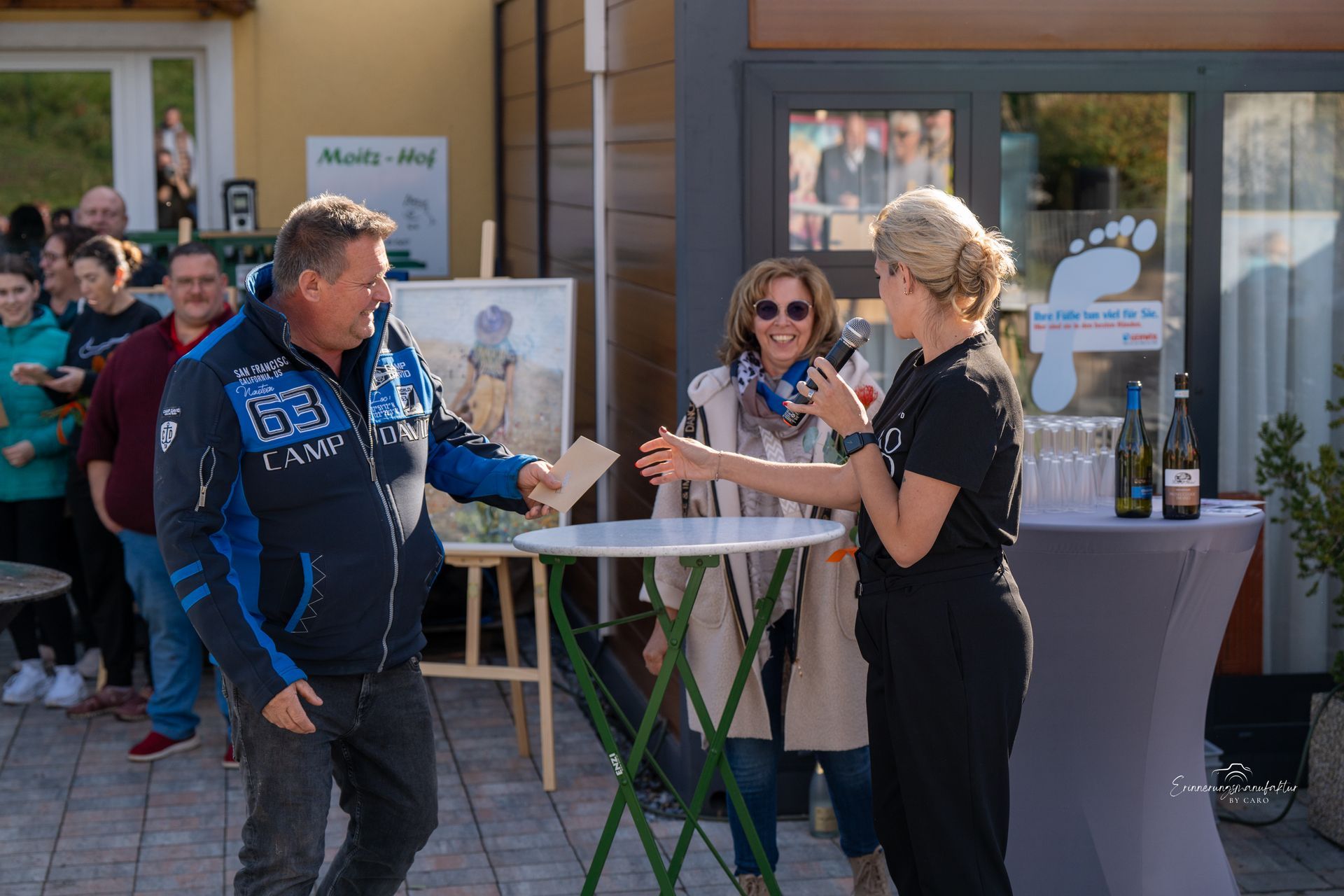 Ein Mann erhält bei einer Freiluftveranstaltung von einer Frau ein Dokument. Umstehende beobachten das Geschehen.