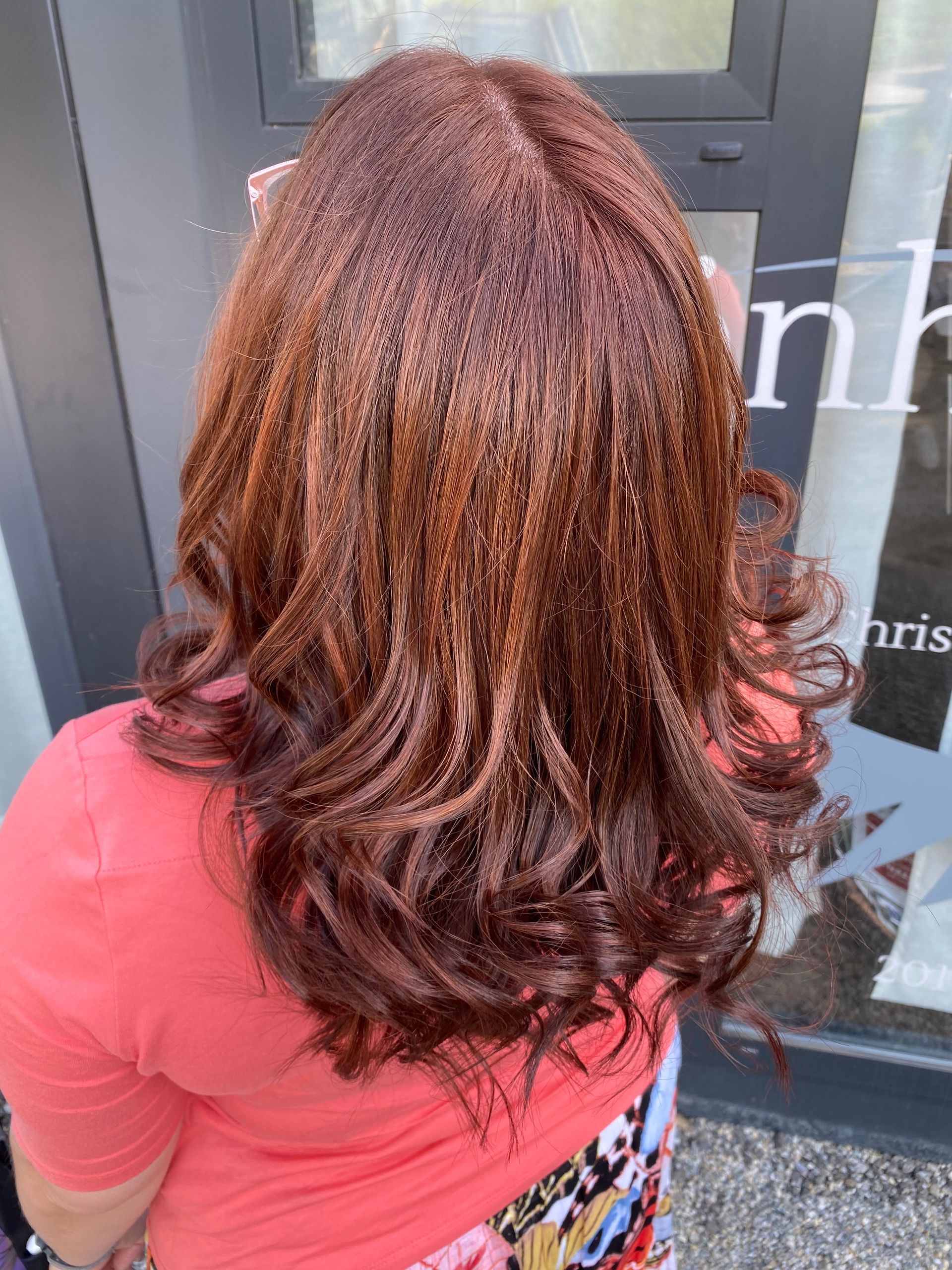 Eine Frau mit rötlich-braunem, gewelltem Haar steht vor einem Gebäude. Sie trägt ein korallenfarbenes Hemd.